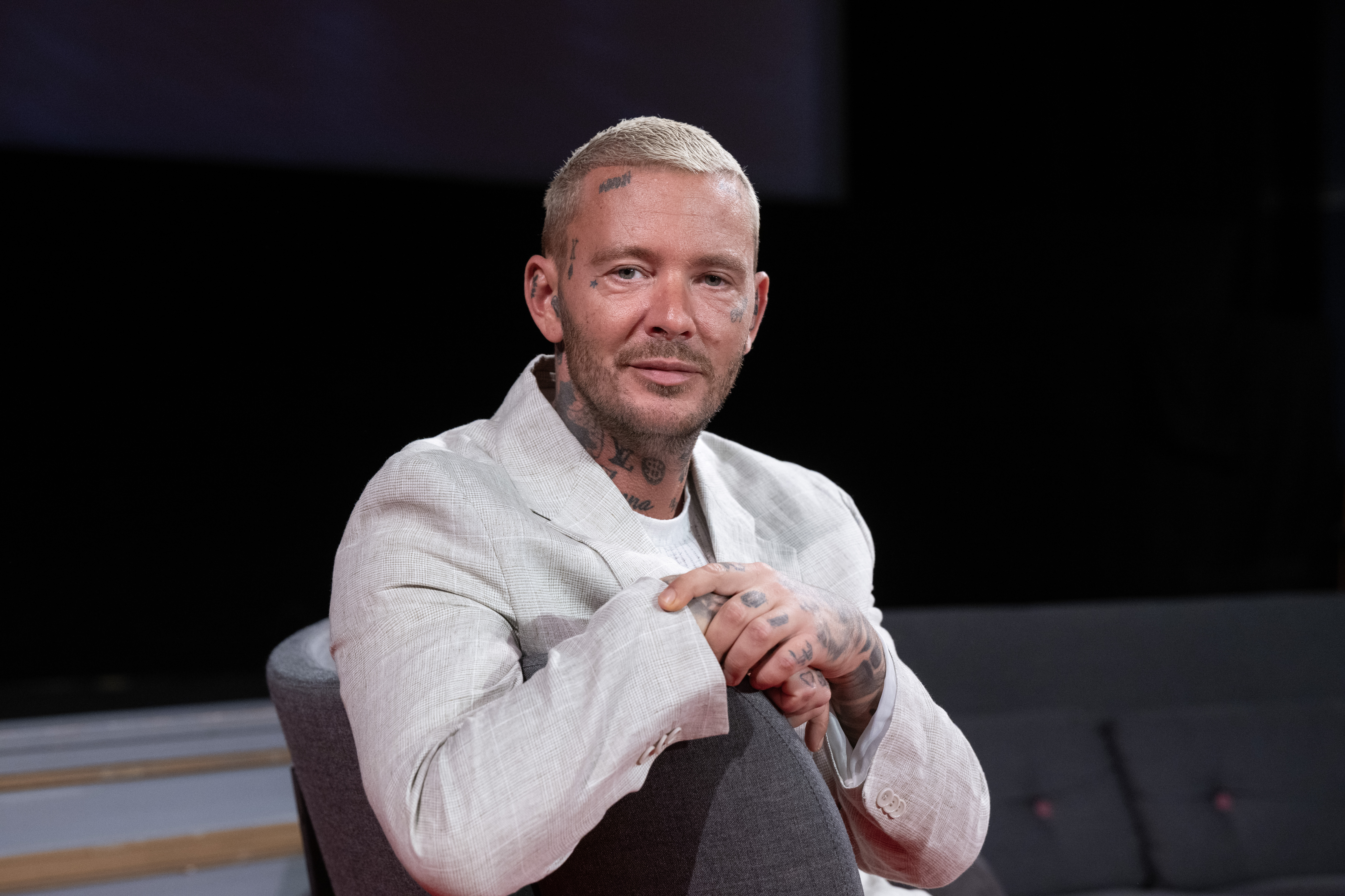 En man med tatueringar i ansiktet och på händerna sitter på en grå stol, bär en ljus kavaj, mot mörk bakgrund.