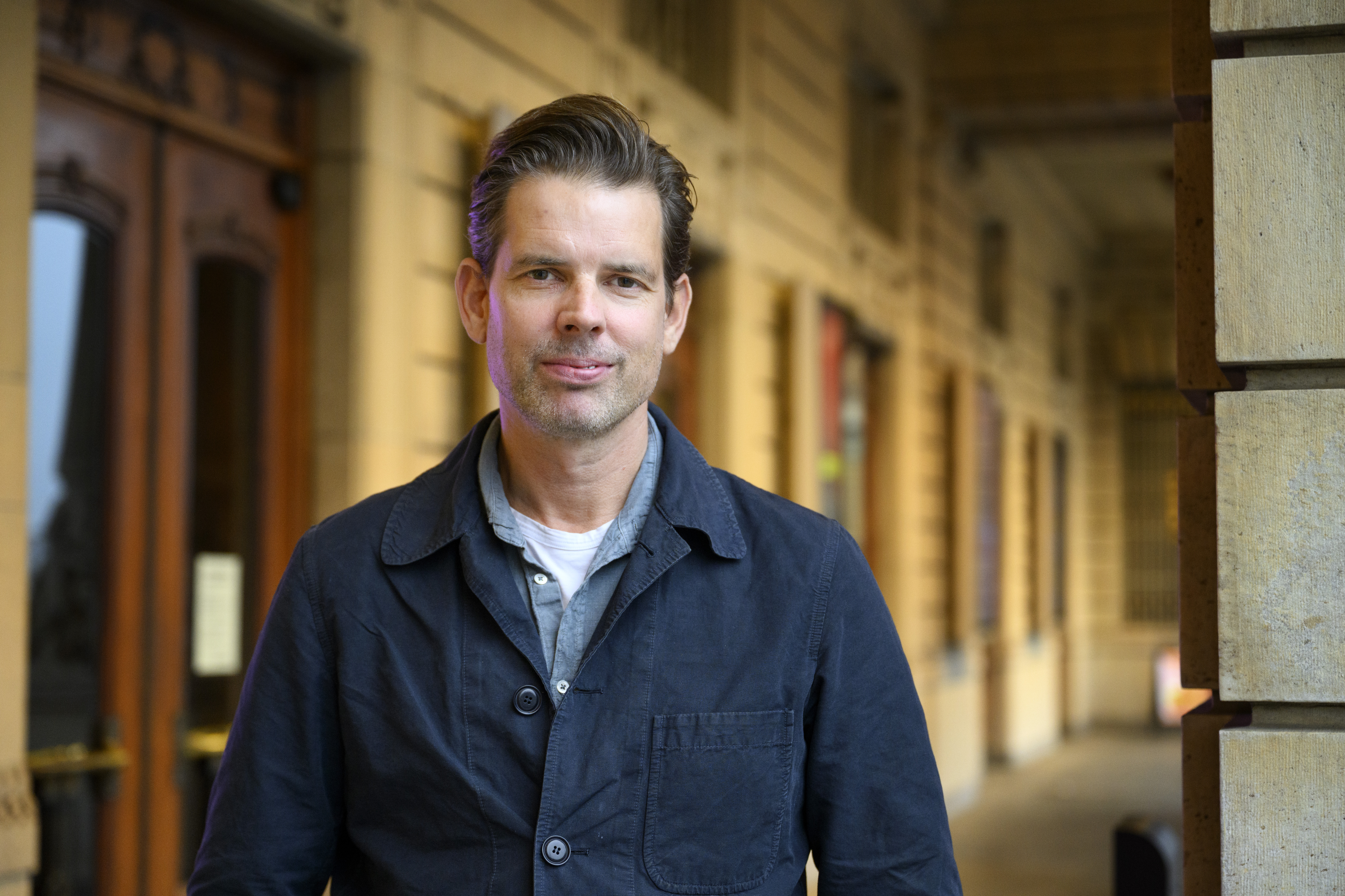 En man med kort brunt hår och mörk jacka över en skjorta står utanför en byggnad med stenfasad och trädörrar.
