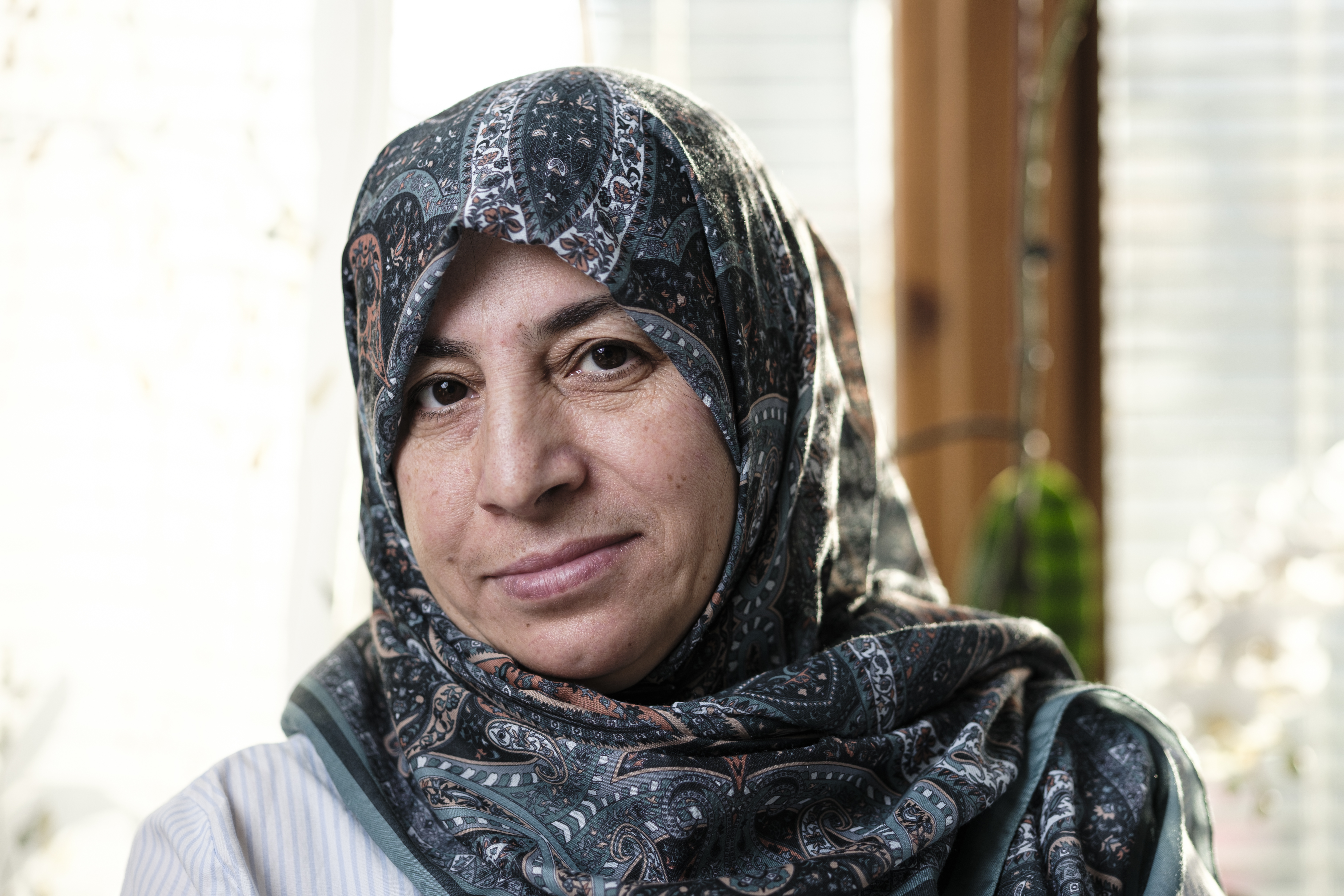 En kvinna med ett mönstrat huvudscarf tittar in i kameran och ler svagt; miljö inomhus med dagsljus i bakgrunden.