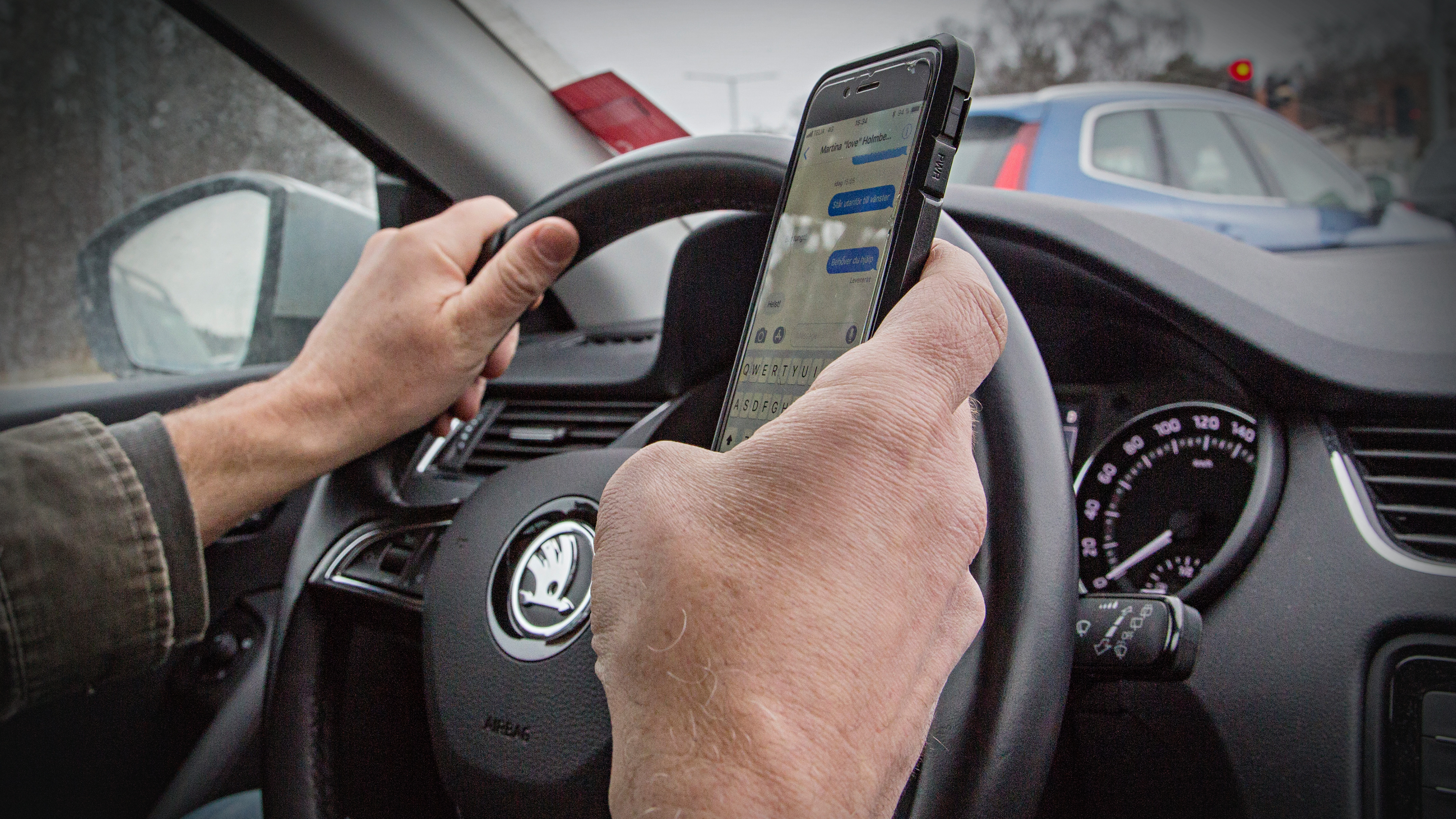 En person kör bil med ena handen på ratten och den andra håller i en mobiltelefon och skriver ett textmeddelande.