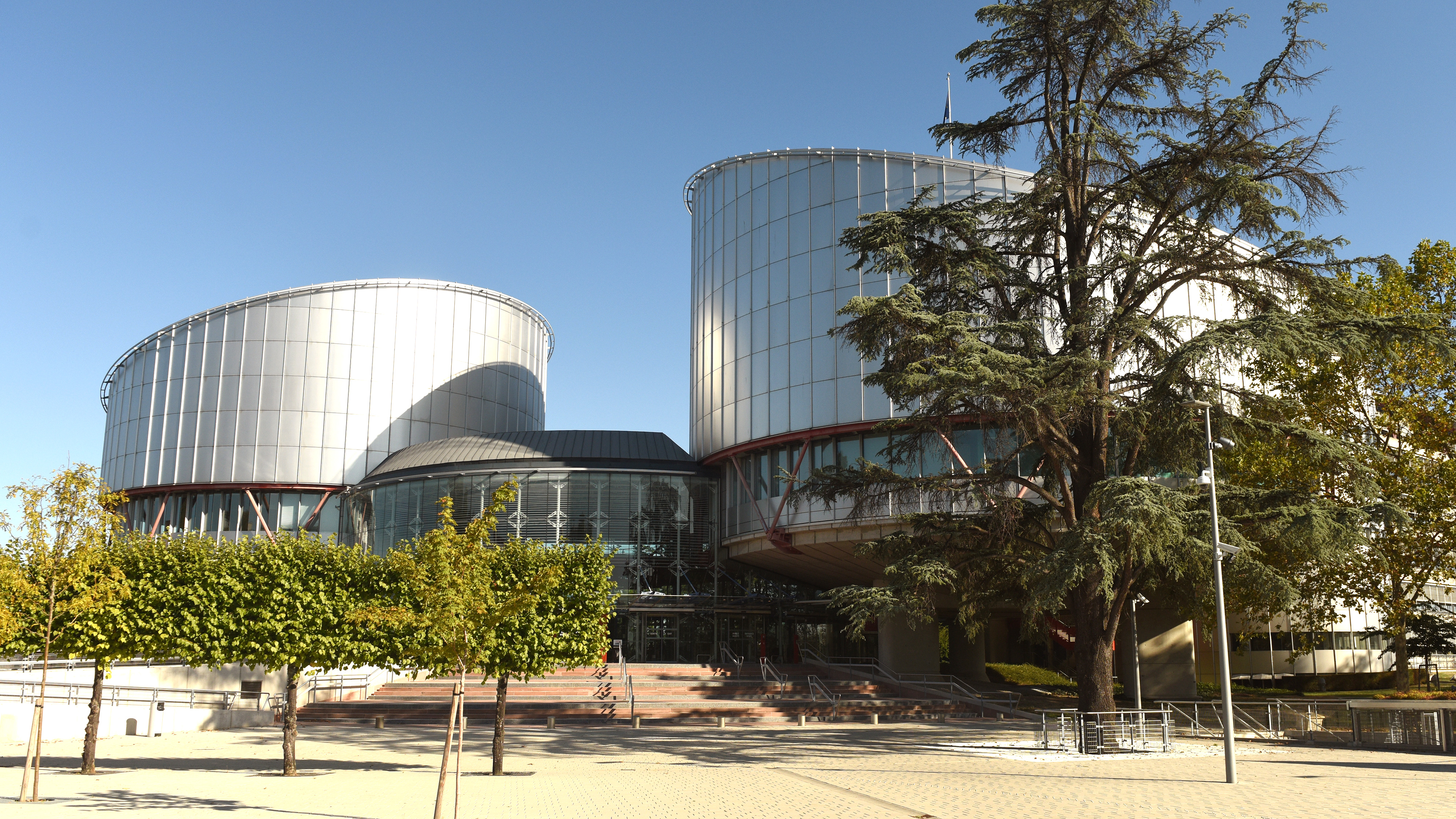 Byggnad av glas och stål med två cylindriska torn, omgiven av träd, med himlen synlig ovanför.