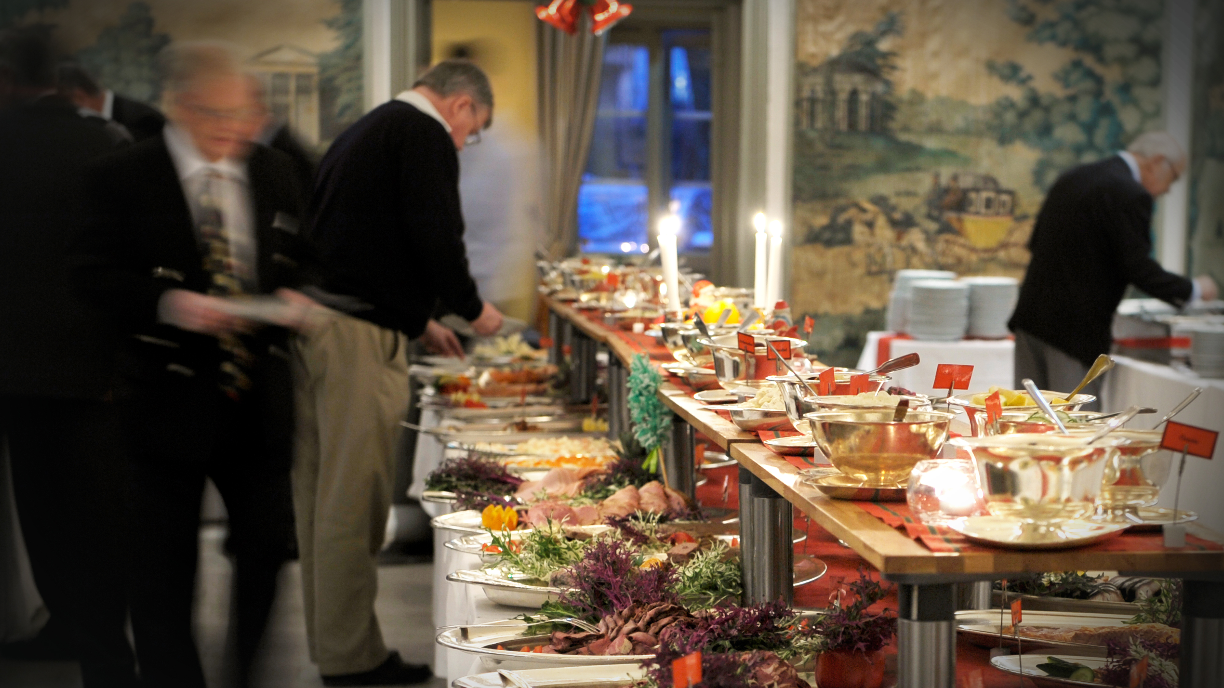 Personer tar mat från ett långt buffébord med olika rätter i en inomhusmiljö med bra belysning.