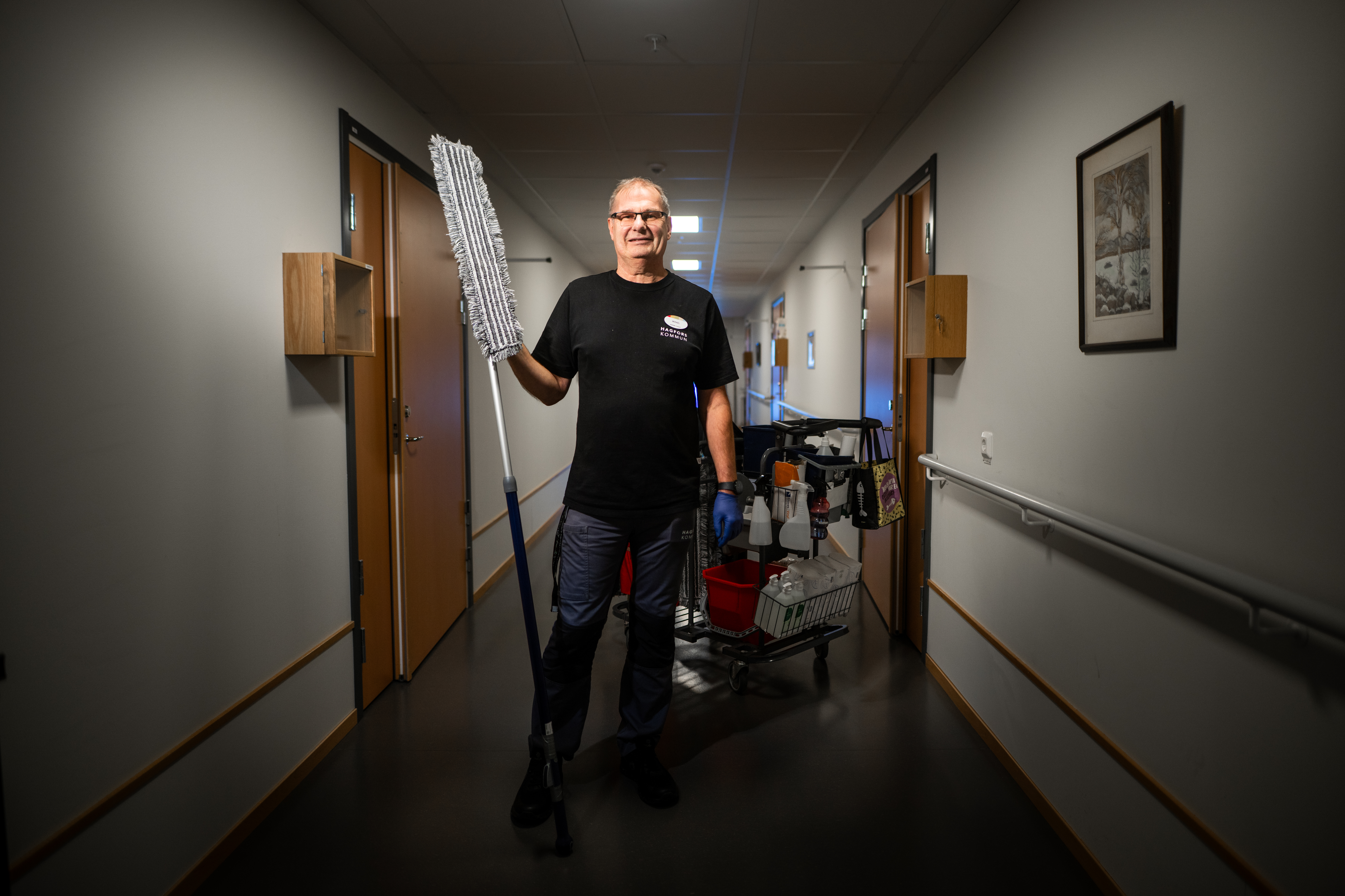 En fastighetsskötare står i en korridor och håller lampkupor, med en städvagn och redskap bakom sig.