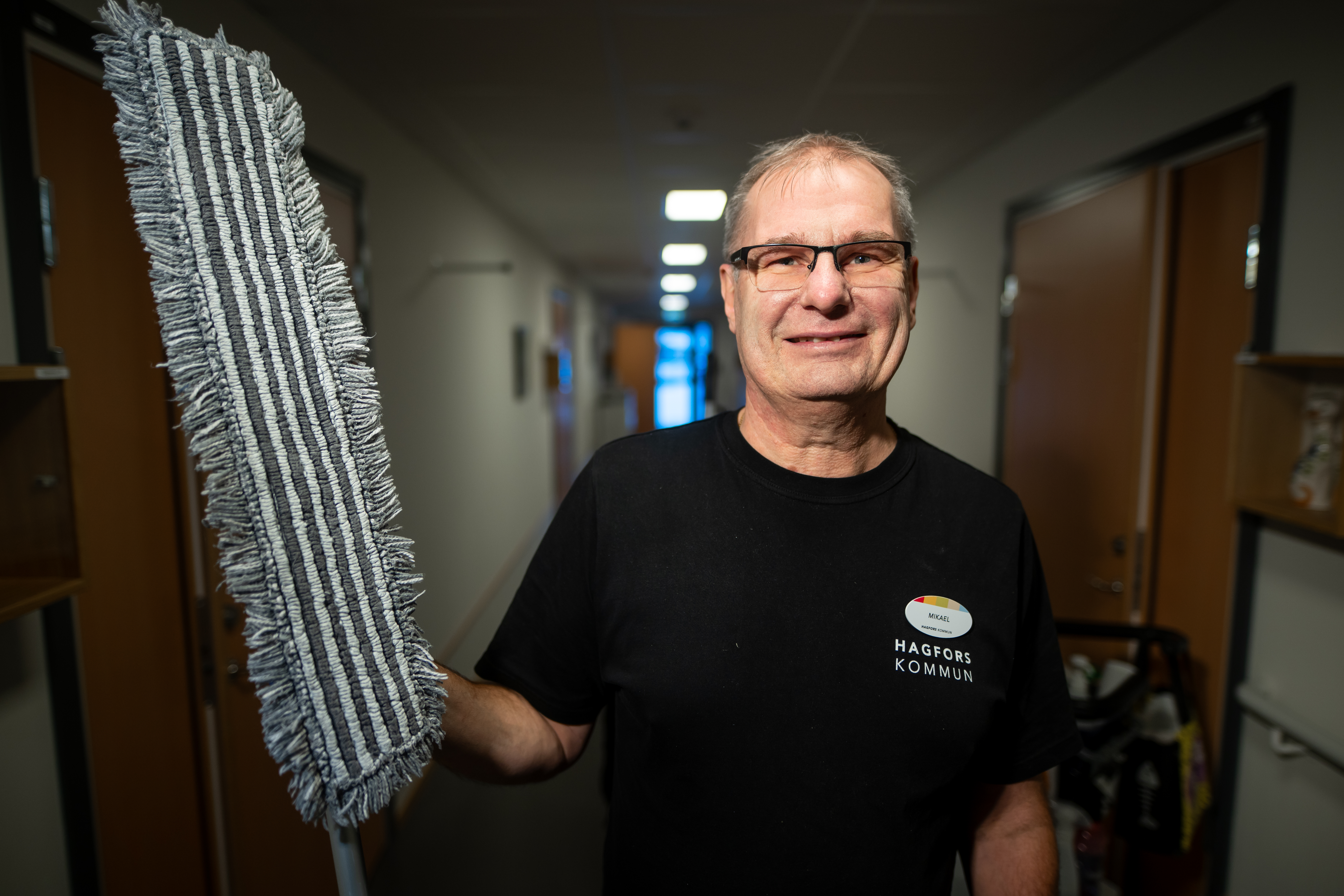 En man med glasögon och en tröja med texten "Hagfors Kommun" står i en korridor och håller i en randig städmopp.