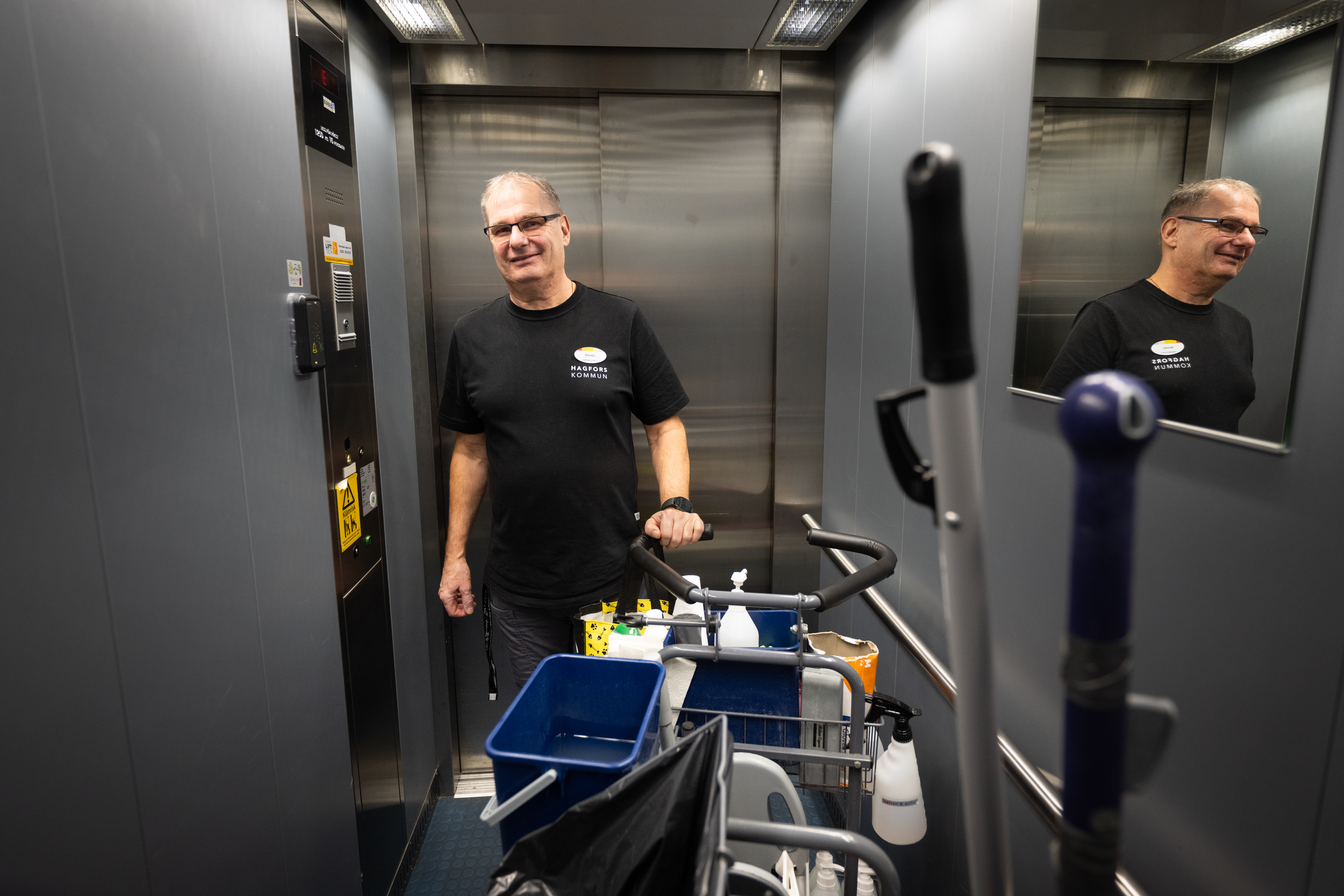 En man i svart skjorta står i en hiss med en städvagn med rengöringsartiklar; hans spegelbild syns i spegeln.