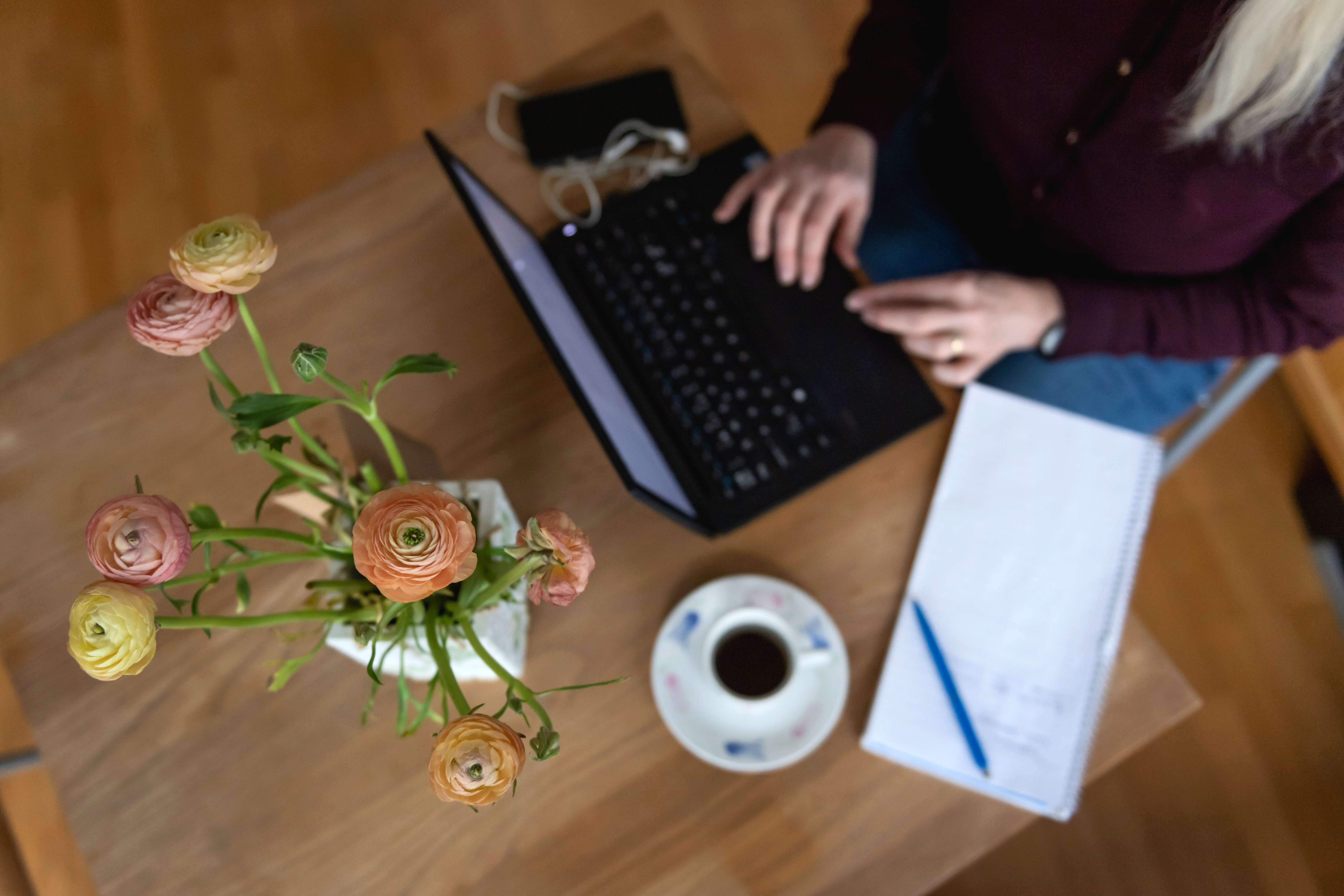 En person skriver på en bärbar dator vid ett bord med en kopp kaffe, ett block med penna, en telefon och en vas med blommor.