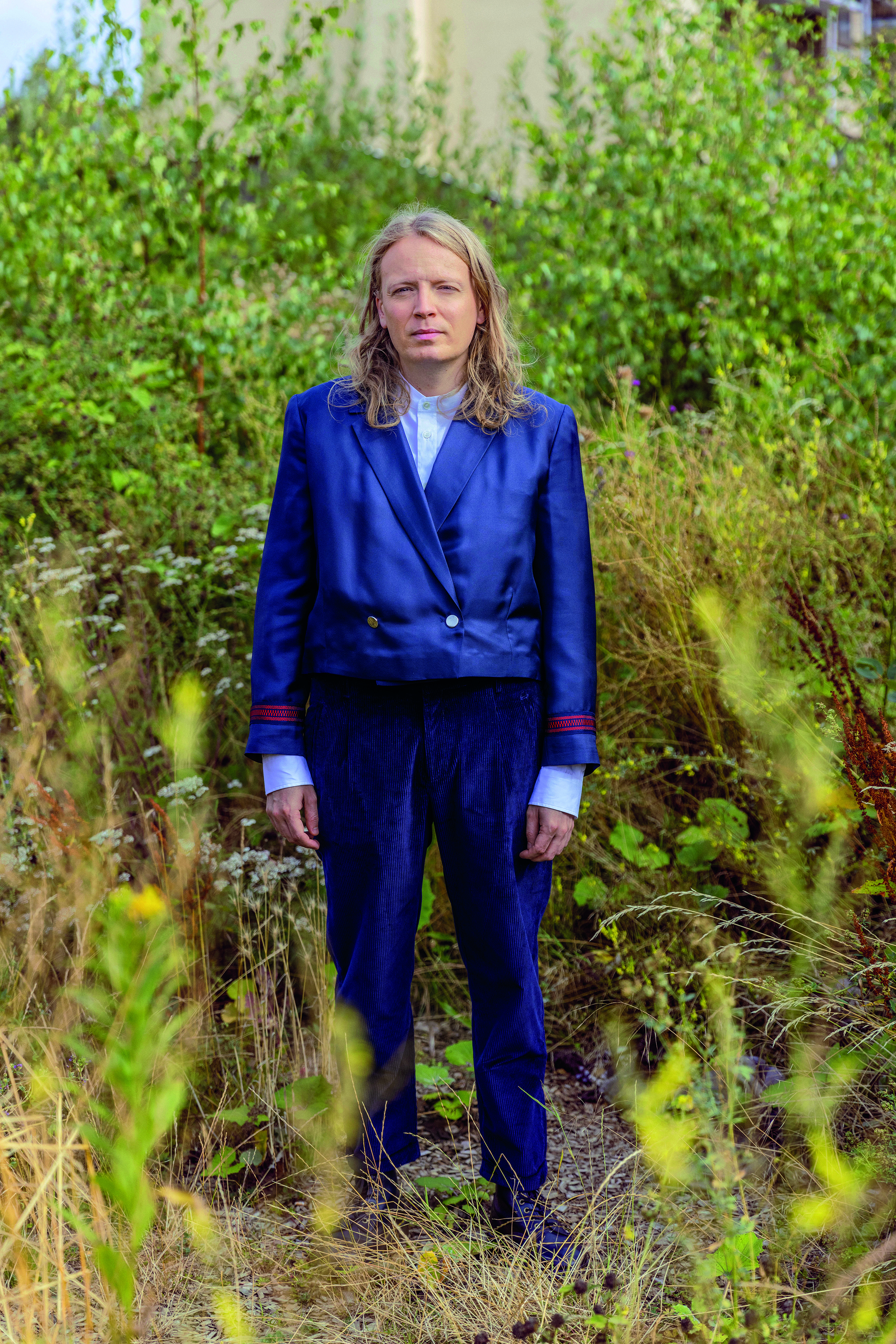 En person med långt blont hår har på sig en marinblå kavaj och byxor och står i ett fält med högt gräs och gröna växter.