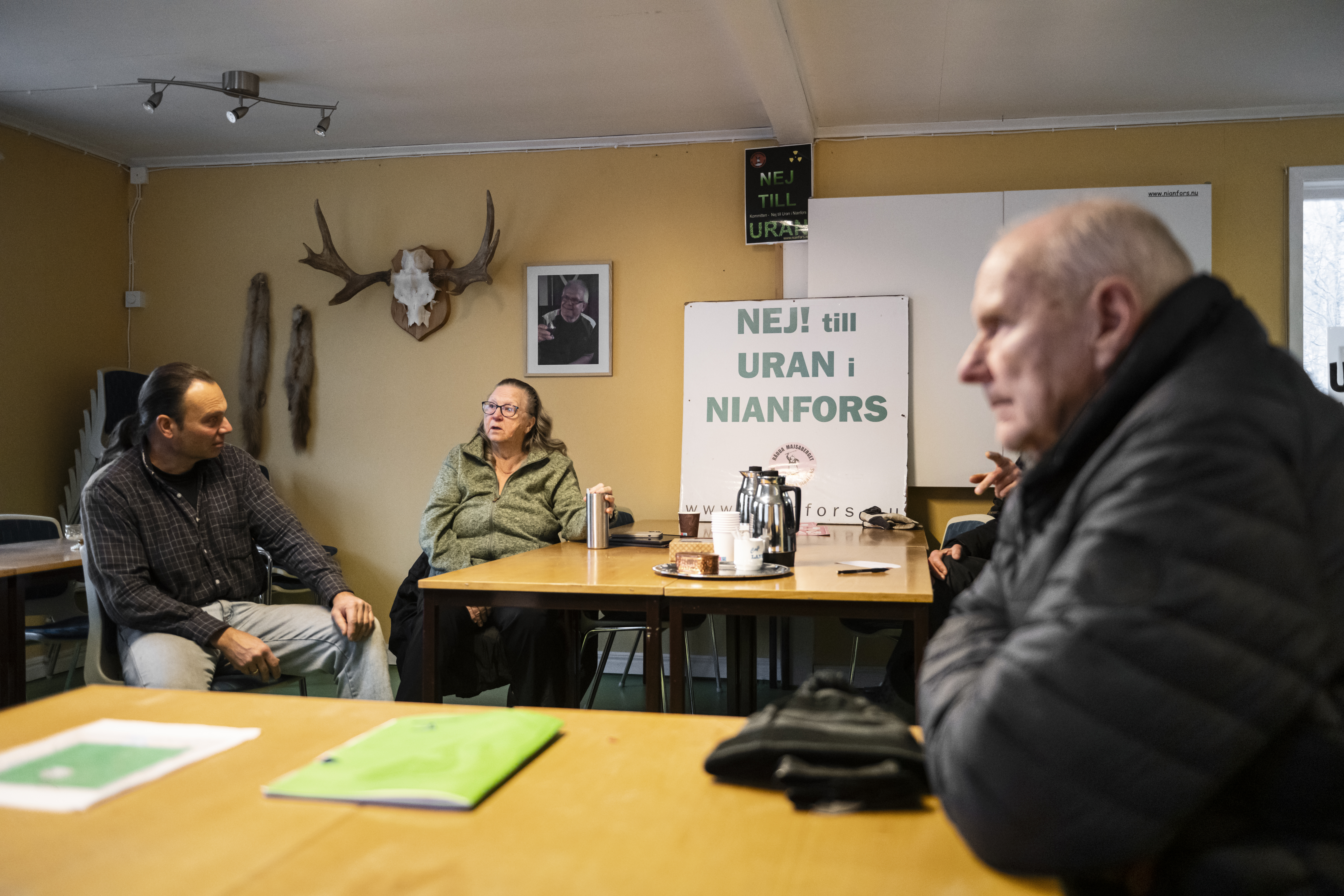Tre personer sitter vid ett bord i ett rum med en skylt där det står "NEJ! till URAN i NIANFORS" och djurskallar på väggen.