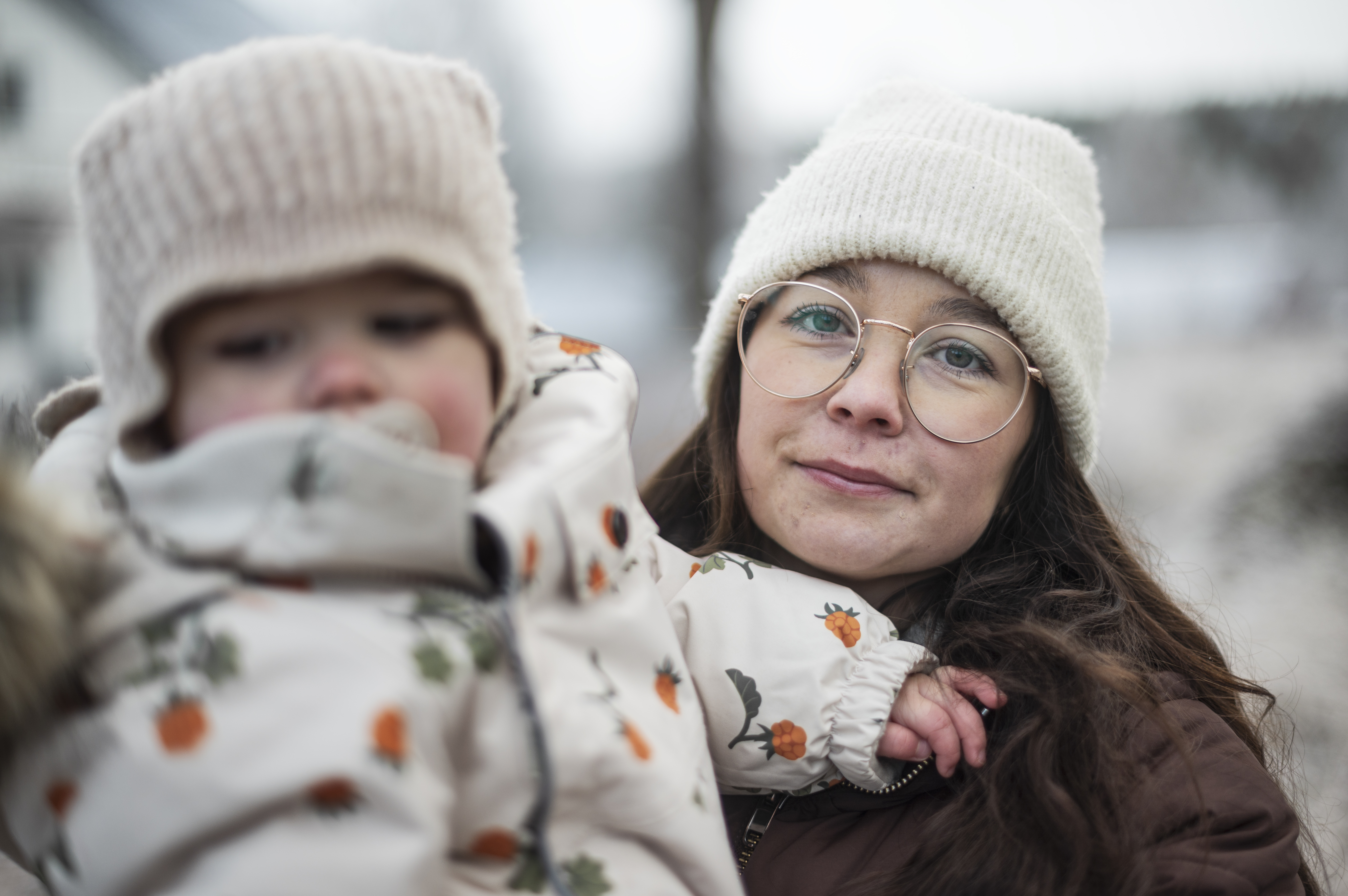 Josefin Wachle med glasögon och vit stickad mössa håller dottern Elise i mönstrade vinterkläder och beige mössa utomhus på vintern.