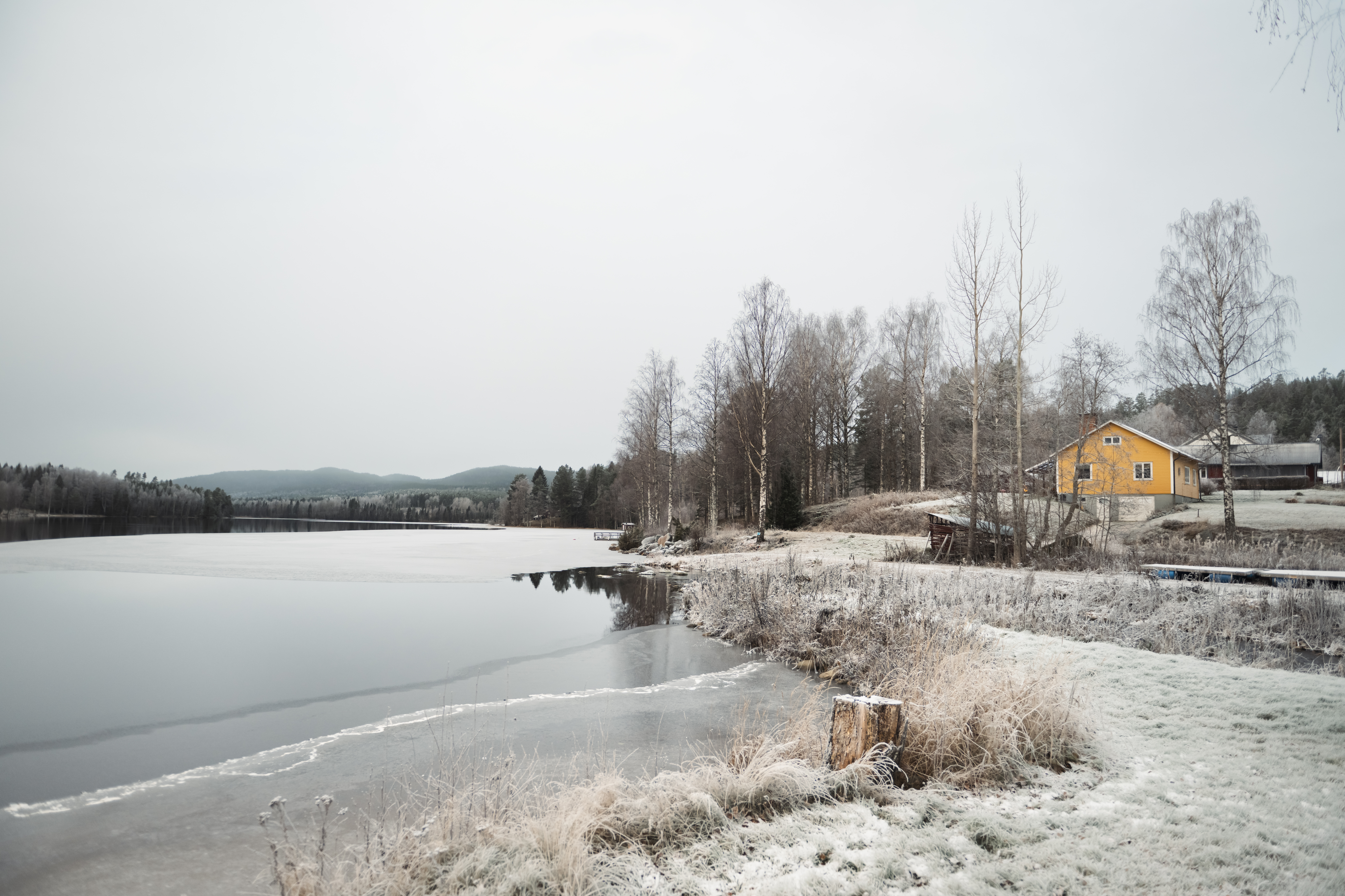 Ett gult hus står vid en delvis frusen sjö med träd utan löv och gräs täckt av frost under en mulen dag.