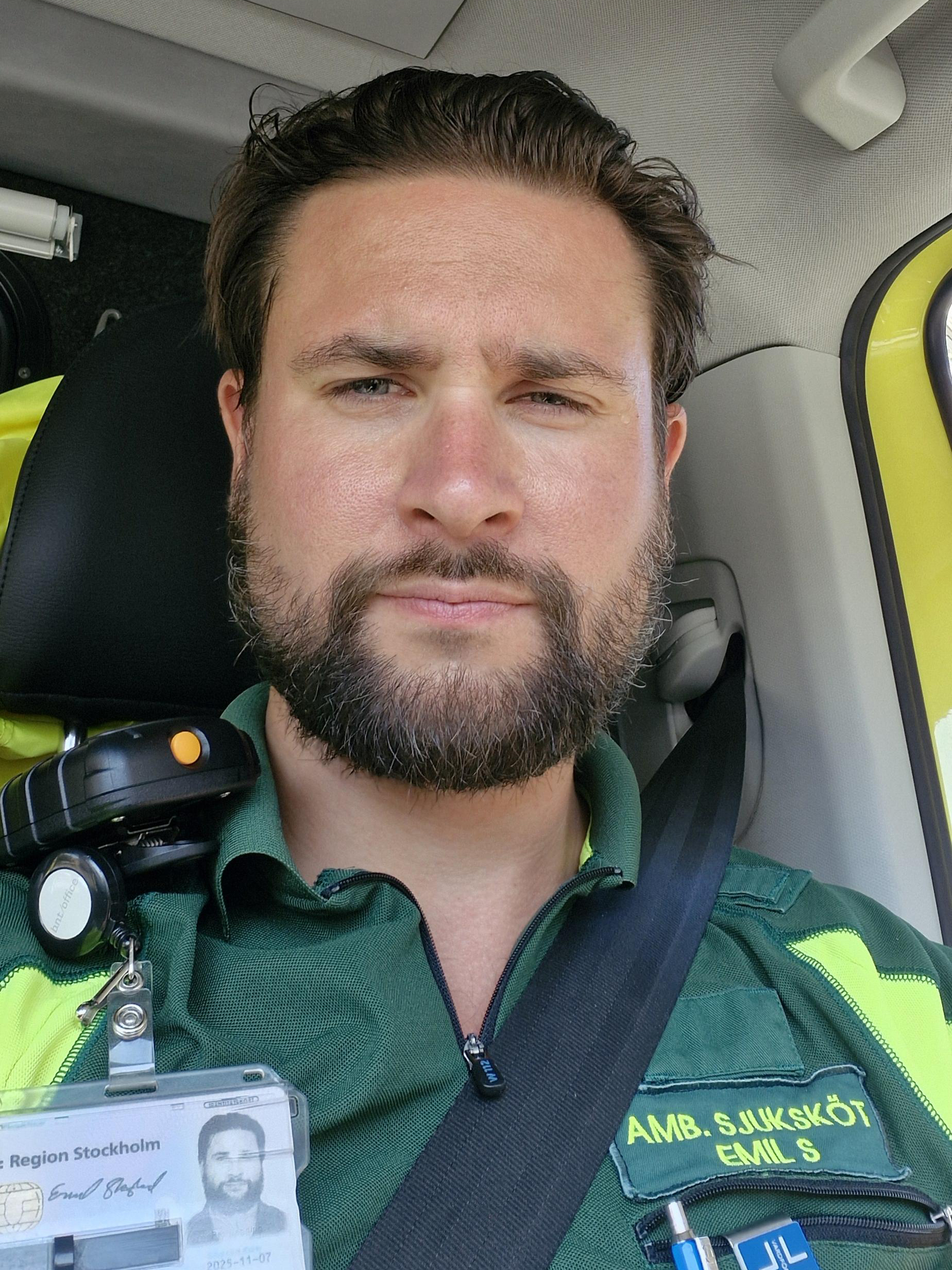 Emil Skoglund i grön sjukvårdsuniform sitter i ett fordon med säkerhetsbälte och har en ID-bricka på skjortan.
