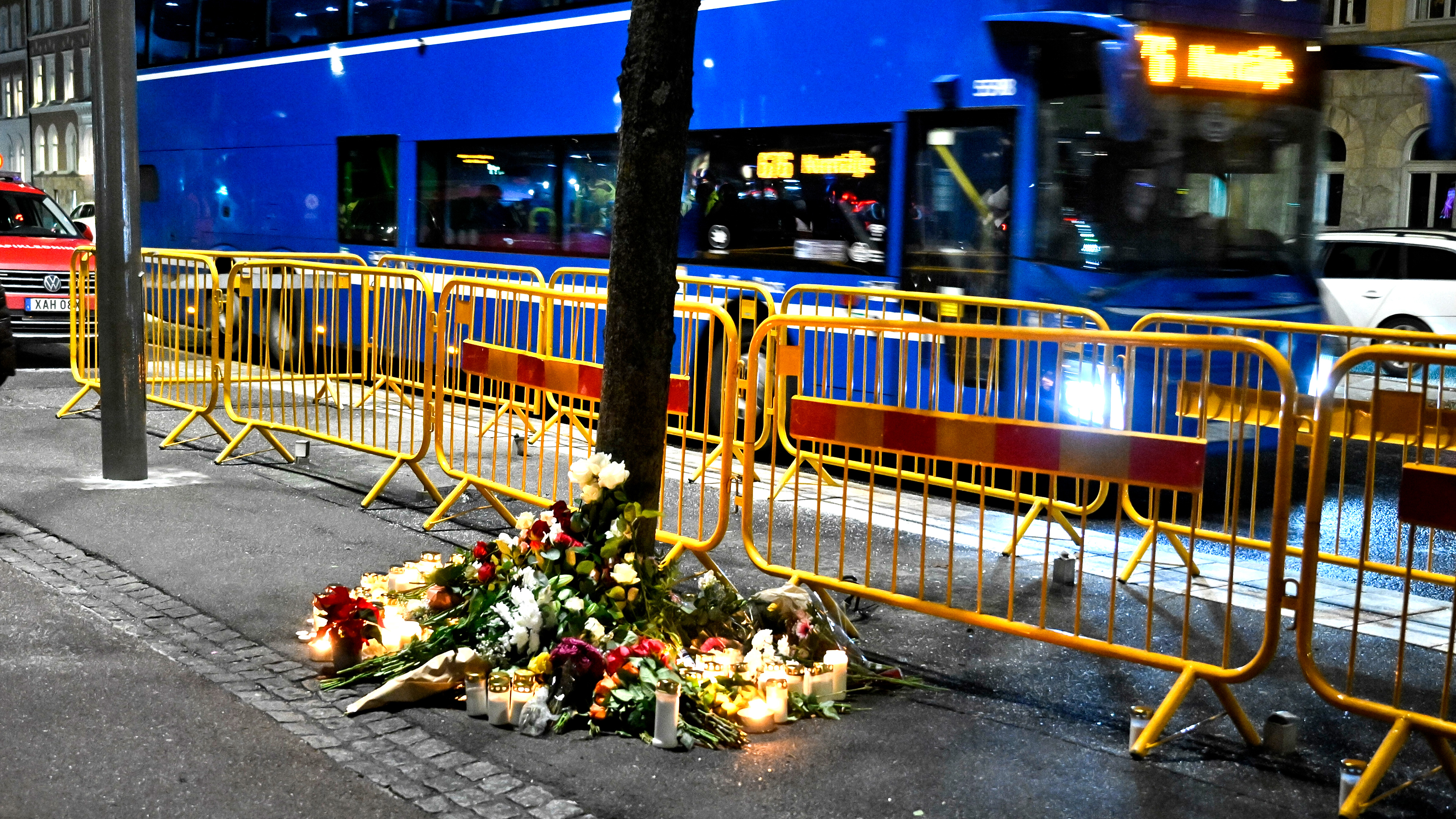 Blommor, ljus och hyllningar ligger vid foten av ett träd på en trottoar, med gula avspärrningar och en blå buss i bakgrunden.