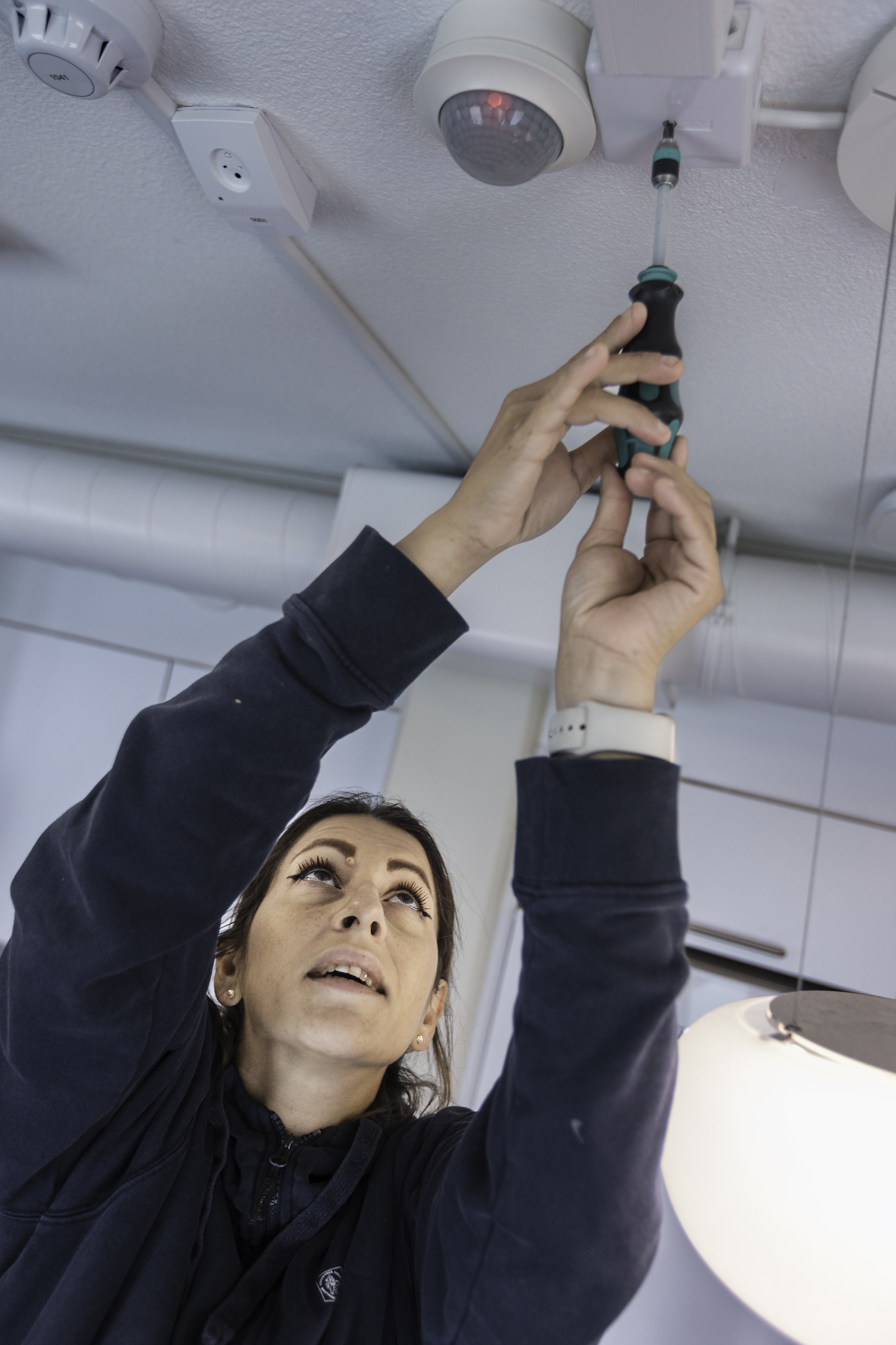 Person använder en skruvmejsel för att montera eller justera en enhet i taket, med kablar och sensorer synliga ovanför.