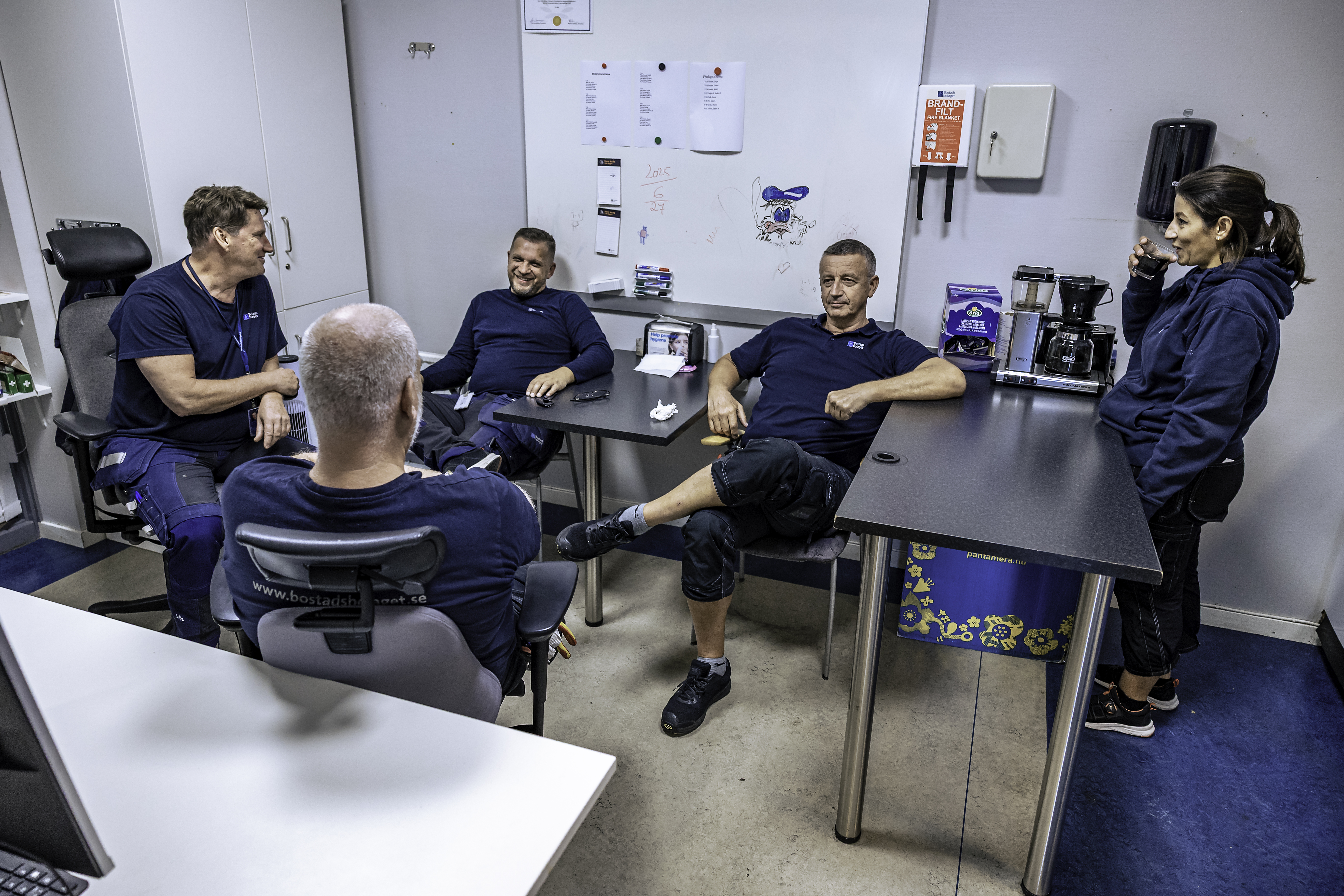 Fem personer i arbetskläder sitter och står runt ett bord i ett pausrum och samtalar. Whiteboard, kaffebryggare och kontorsmaterial syns.