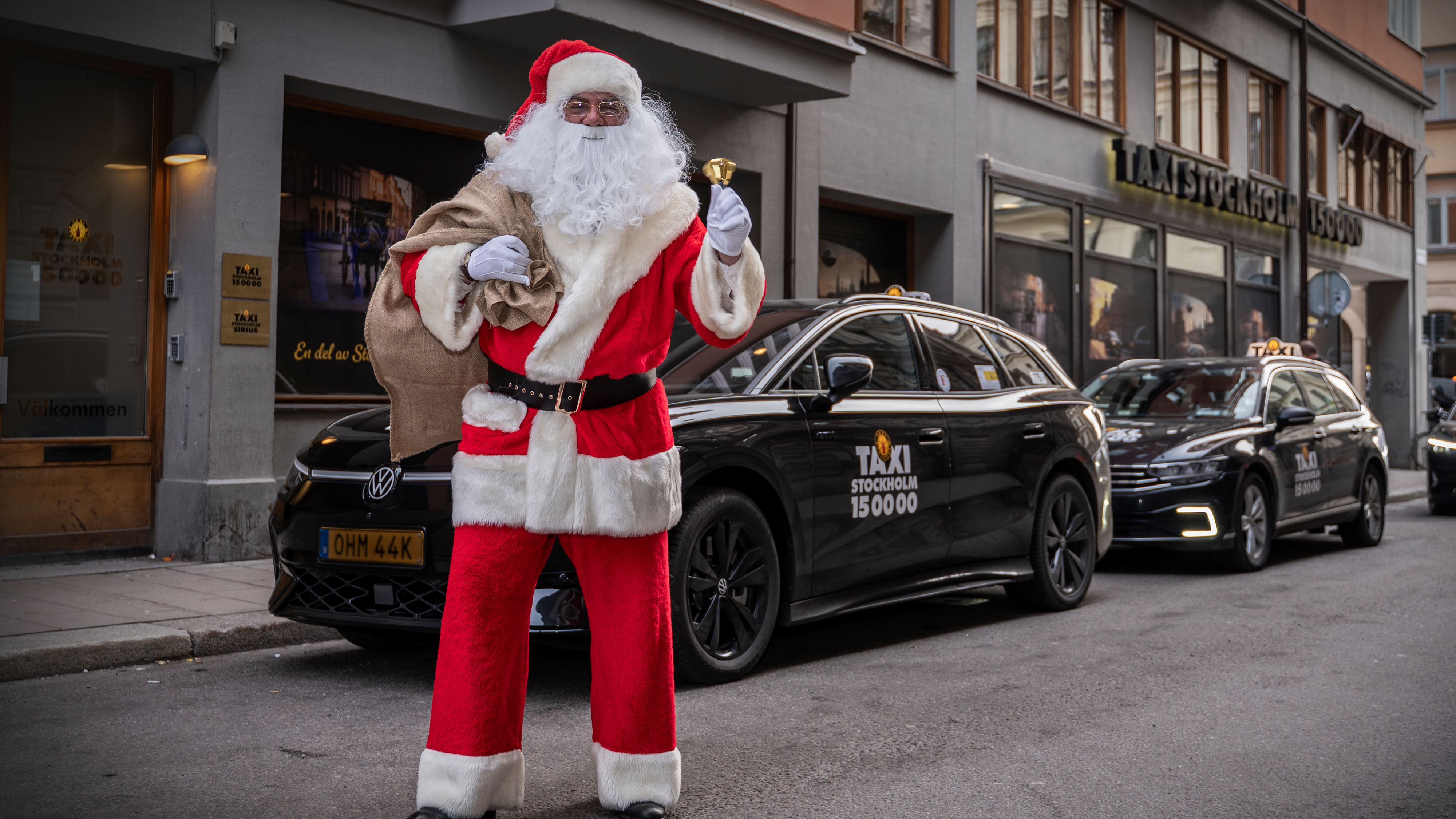 En person utklädd till jultomte står på en stadsgata, håller i en klocka och säckar, med två svarta taxibilar bakom sig.