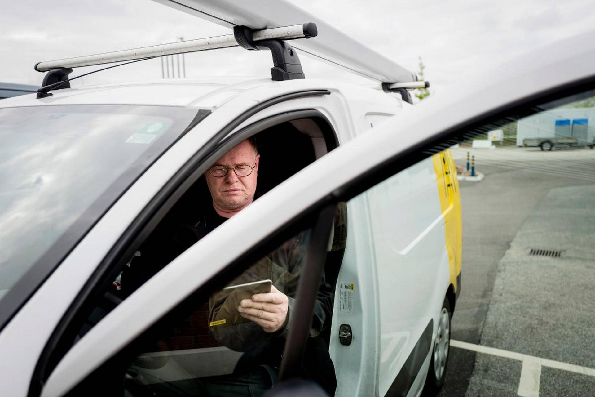 En man sitter i förarsätet i en vit skåpbil och tittar på en surfplatta med dörren öppen på en parkeringsplats.
