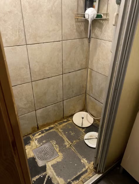 En liten duschkabin med trasiga golvplattor, ett avlopp och en hörnhylla med toalettartiklar samt en vit handdusch.