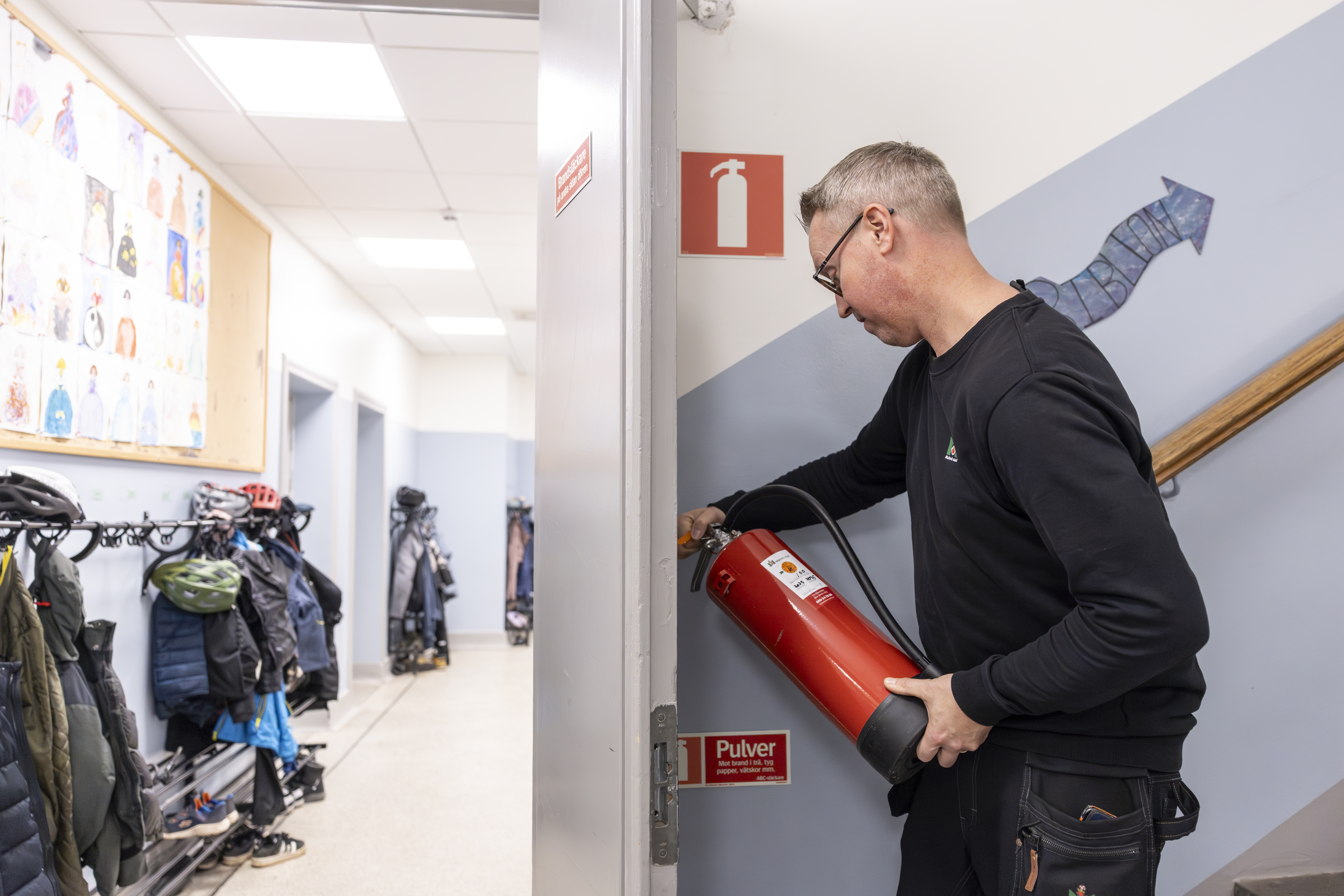 Jesper Sköld sätter tillbaka en brandsläckare i dess väggfäste i en hall med jackor och hjälmar som hänger på krokar.