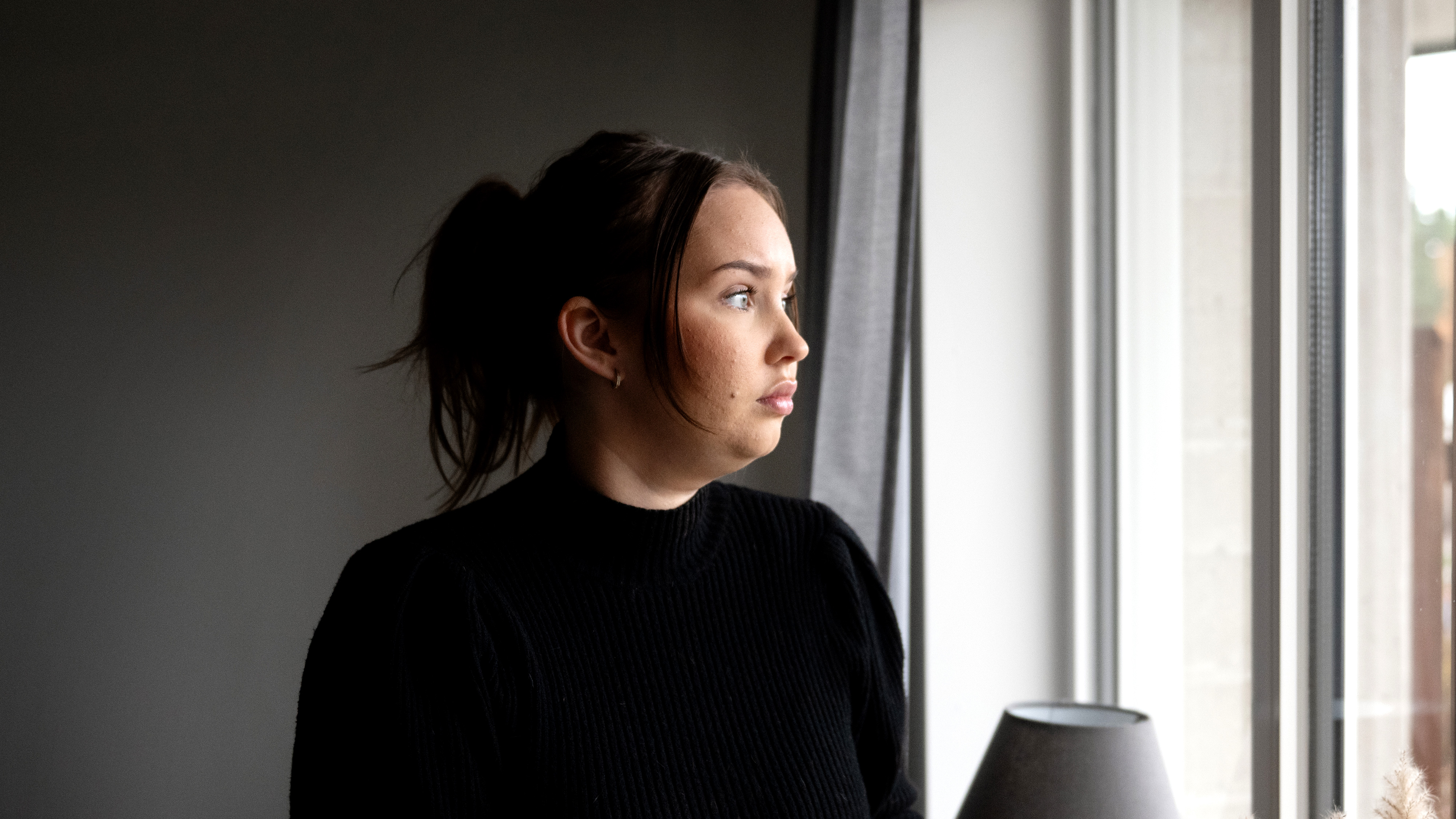 Isabelle Lötell med brunt hår i en hästsvans, iförd en svart tröja, står inomhus och tittar ut genom ett fönster utan att le.