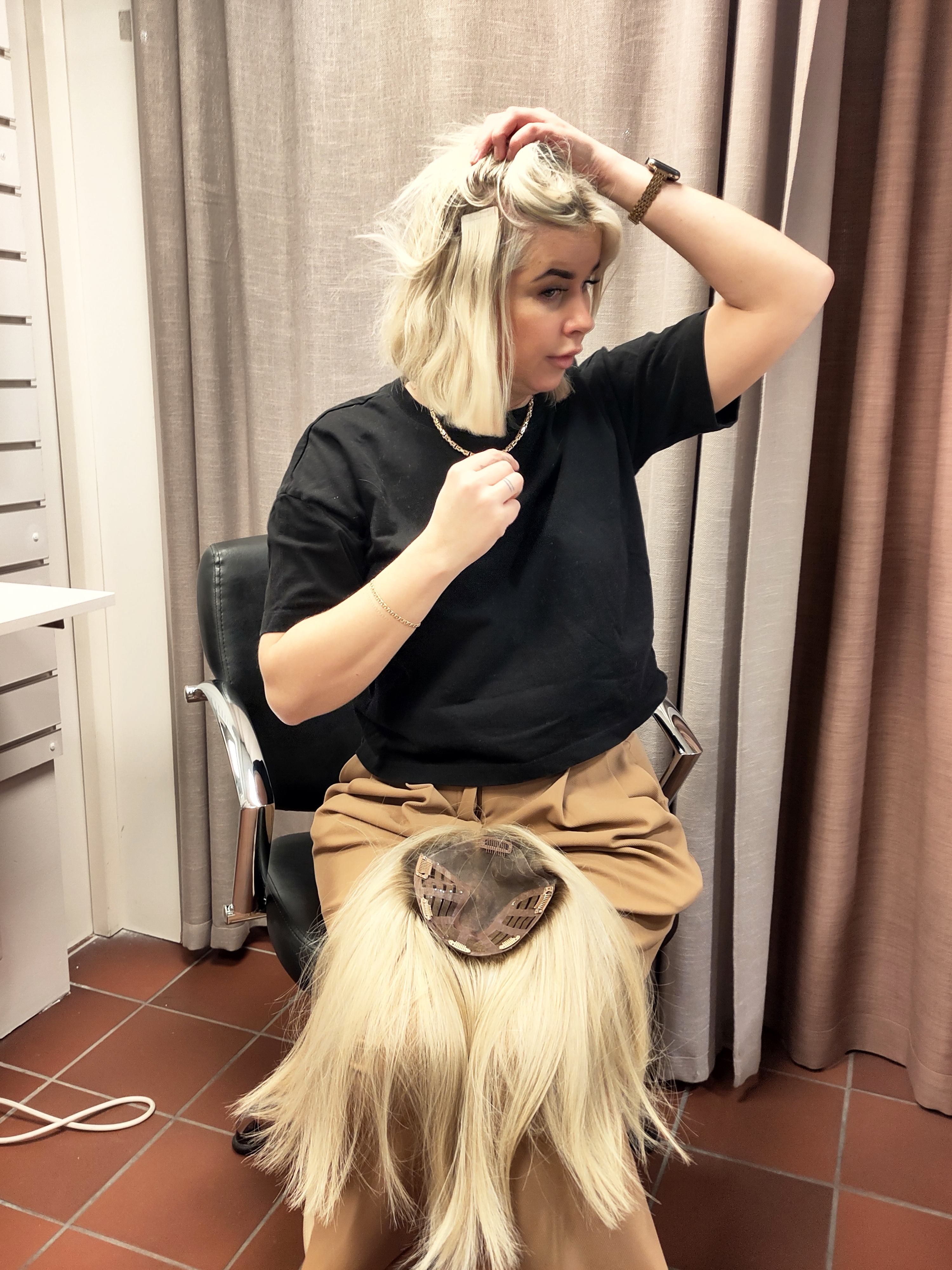 En person med kort blont hår sitter i en frisörstol, håller huvudet och har en lång blond peruk i knät.