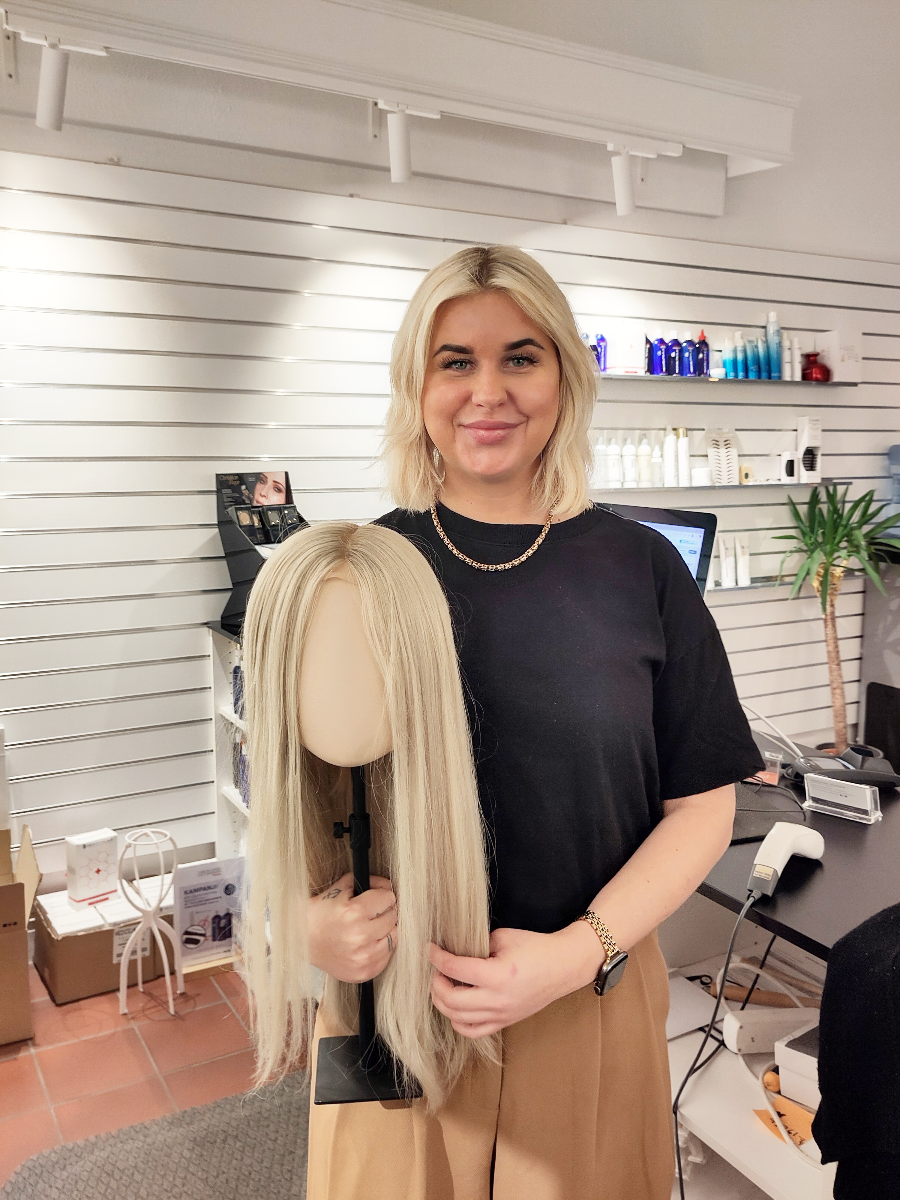 En kvinna med blont hår och svart tröja står inomhus och håller en skyltdockas huvud med långt blont hår framför sig.