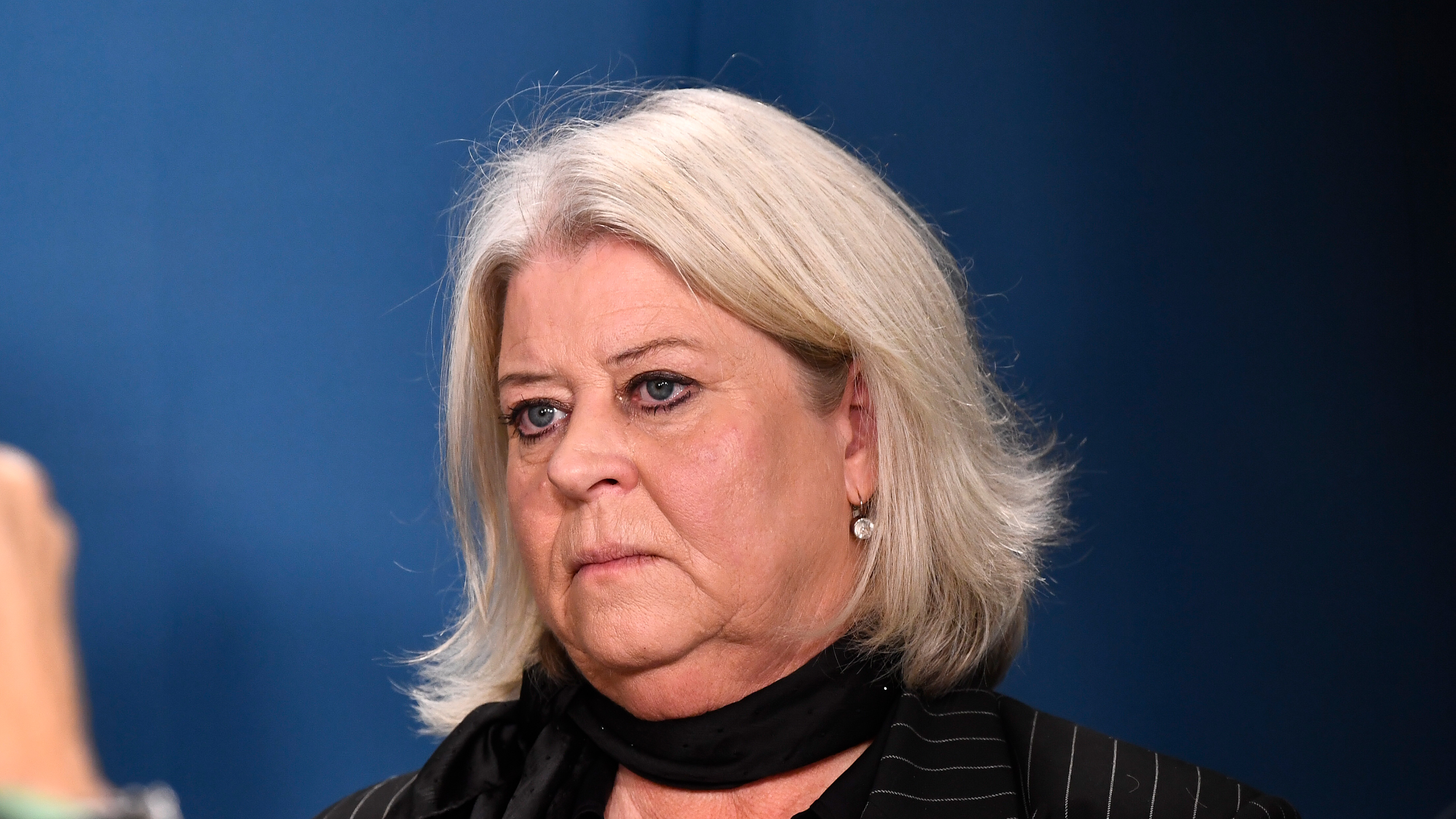 En kvinna med axellångt grått hår och en svart scarf står framför en blå bakgrund och ser allvarlig ut.