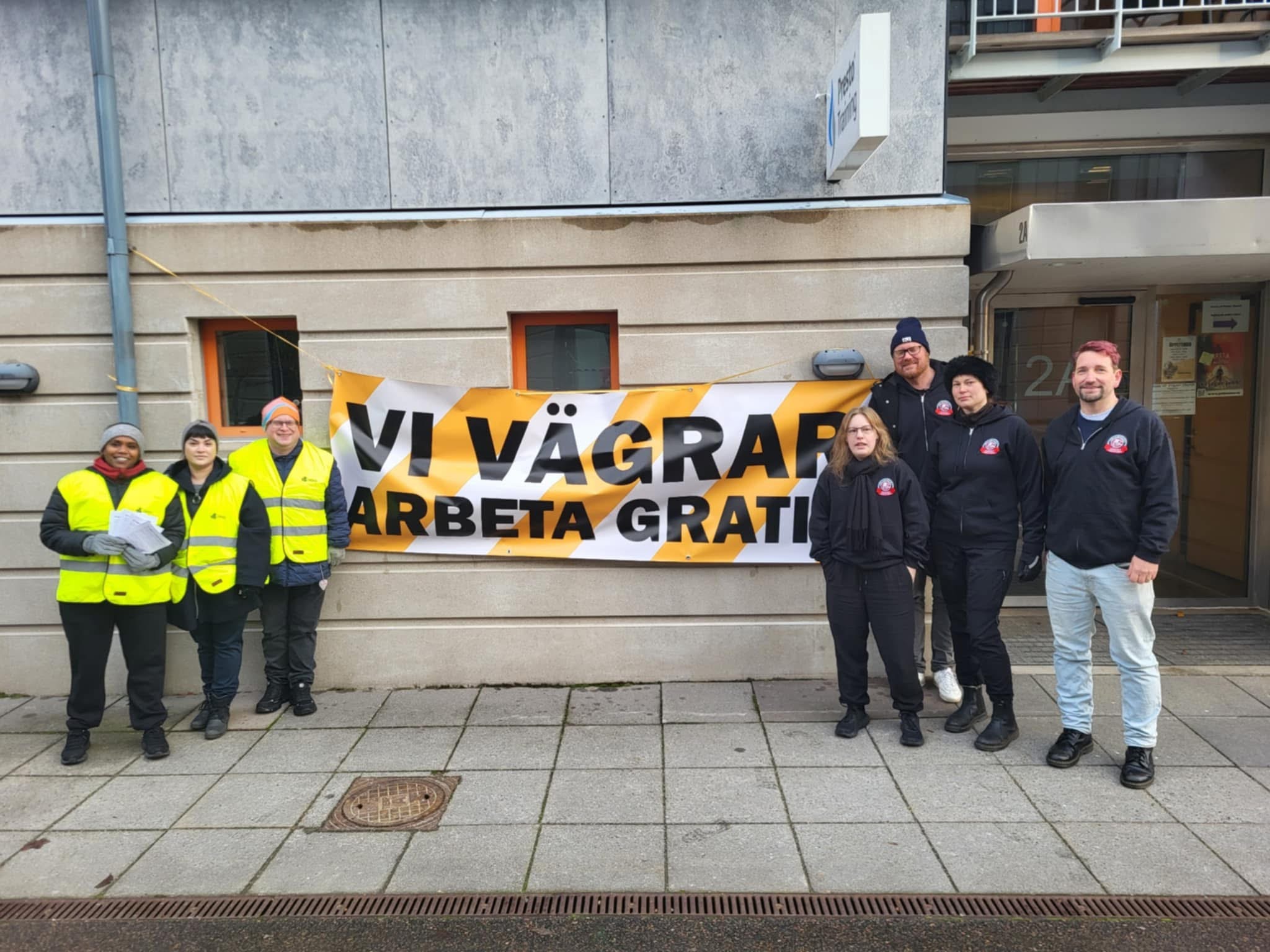 Sju personer står utanför en byggnad bredvid en banderoll med texten "VI VÄGRAR ARBETA GRATIS.