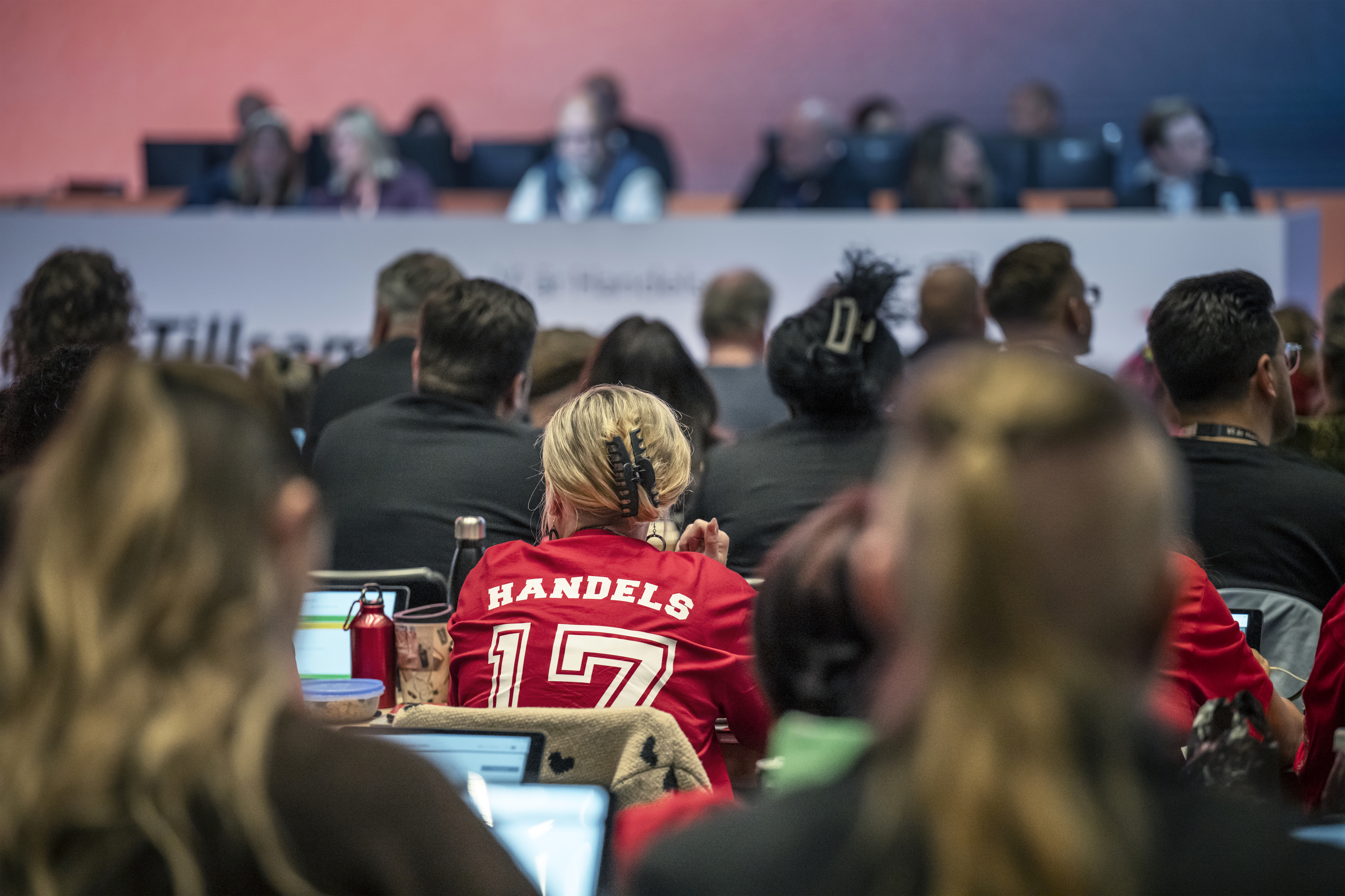 En grupp personer sitter vända mot en panel på en konferens; en person i förgrunden har en röd tröja med "HANDELS 17" på ryggen.