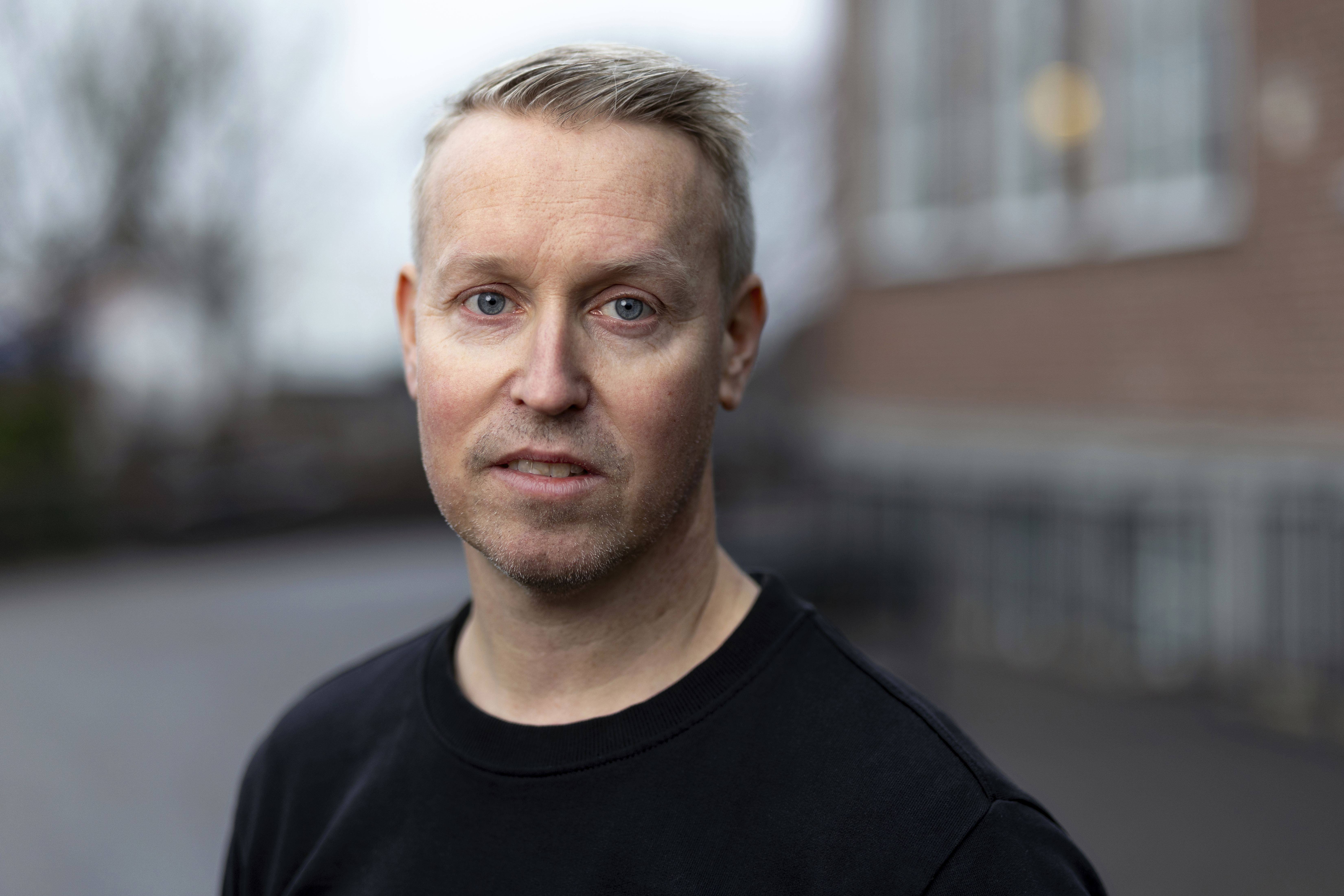 Jesper Sköld står utanför och tittar allvarligt mot kameran. En suddig skolbyggnad skymtar i bakgrunden.