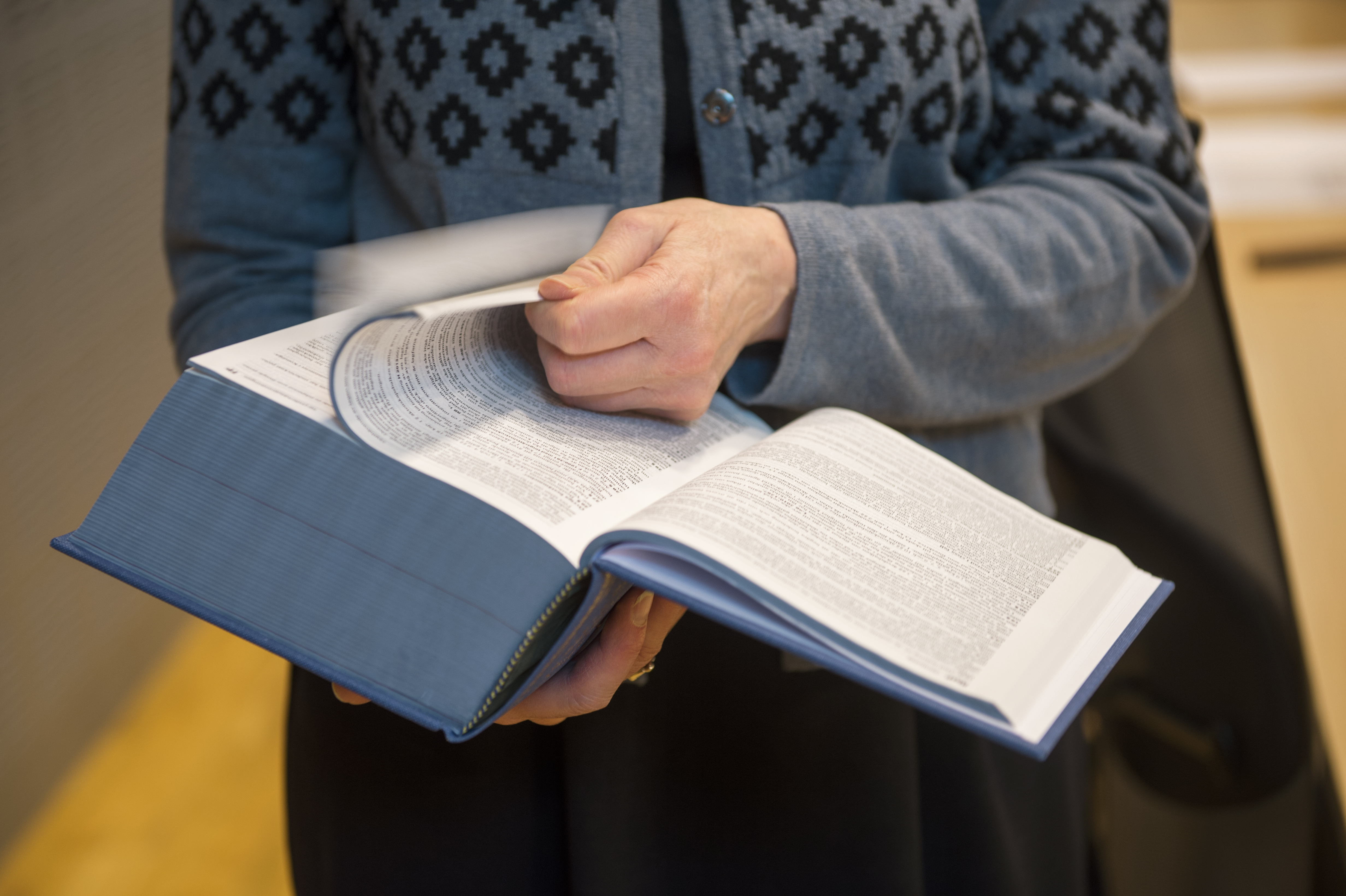 En person i mönsstrad tröja bläddrar i en stor uppslagen bok, kanske ett lexikon eller en faktabok.