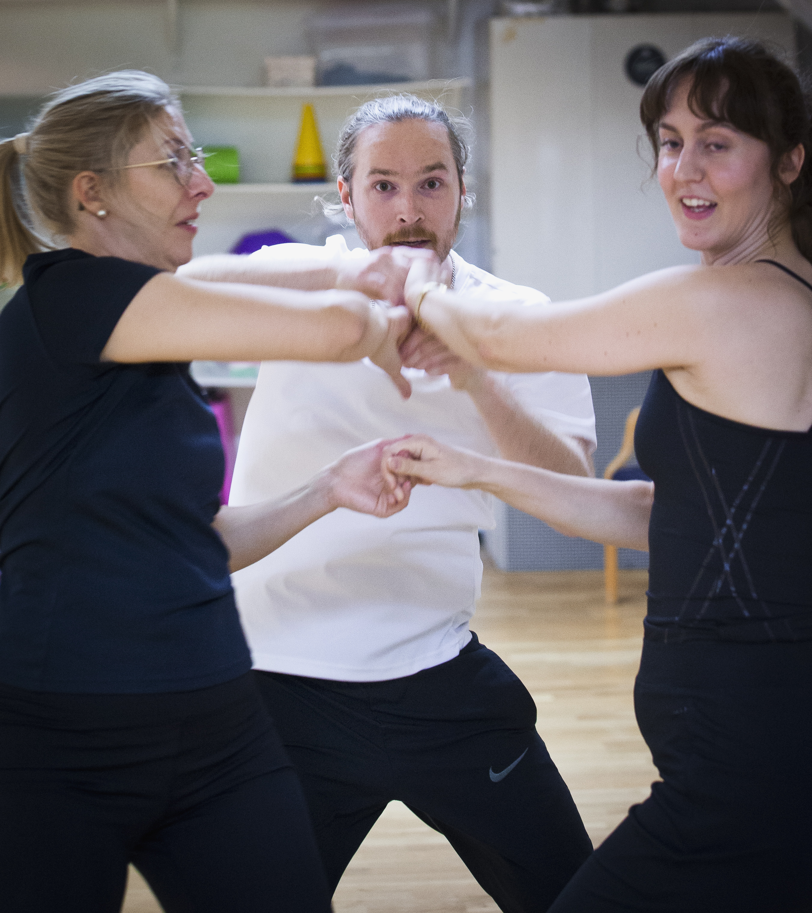 Tre vuxna i träningskläder övar en gruppövning eller dansrutin inomhus och håller varandra om armarna.