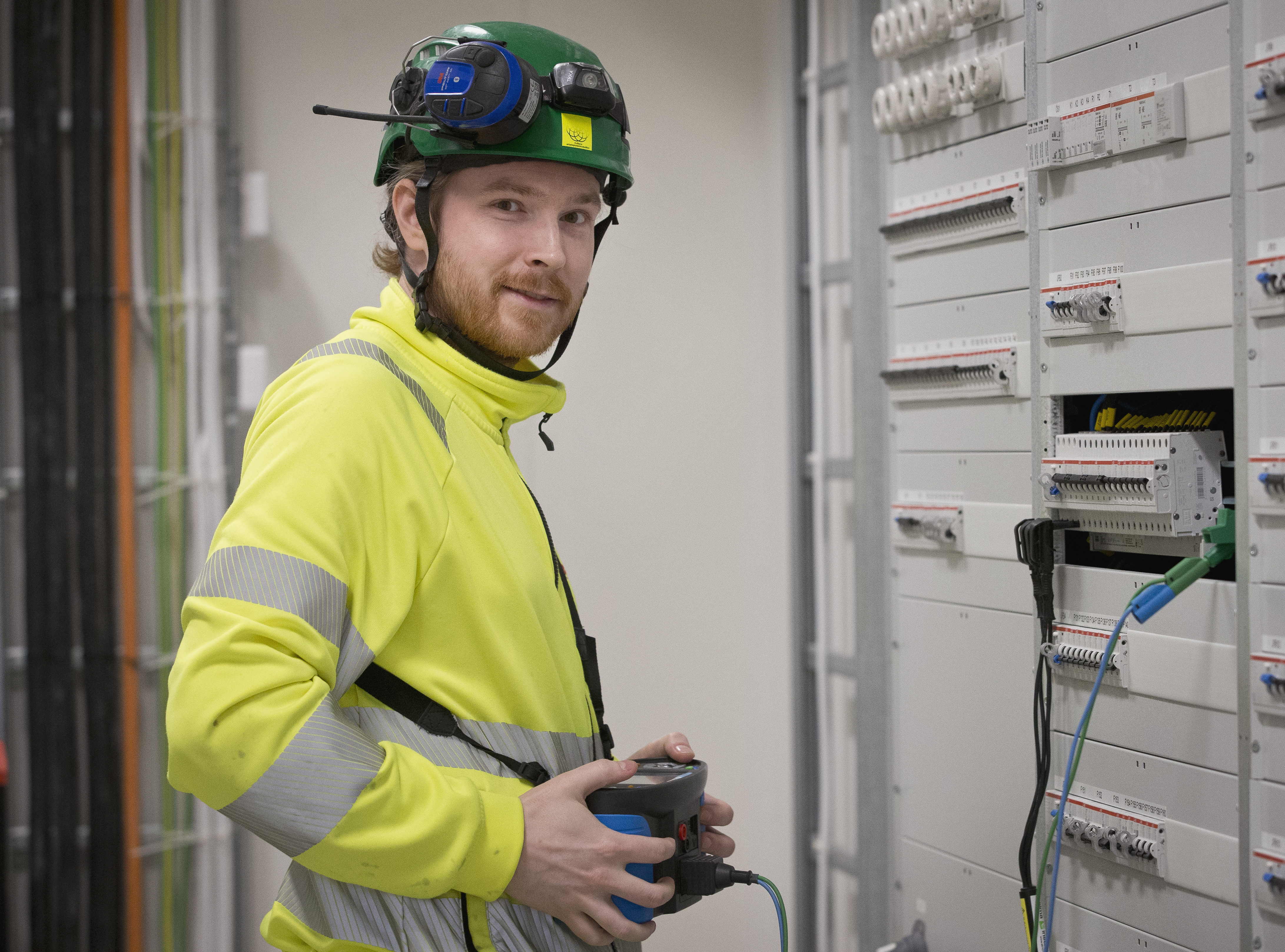 En arbetare med varseljacka och grönt hjälm håller utrustning och står bredvid ett öppet elskåp.