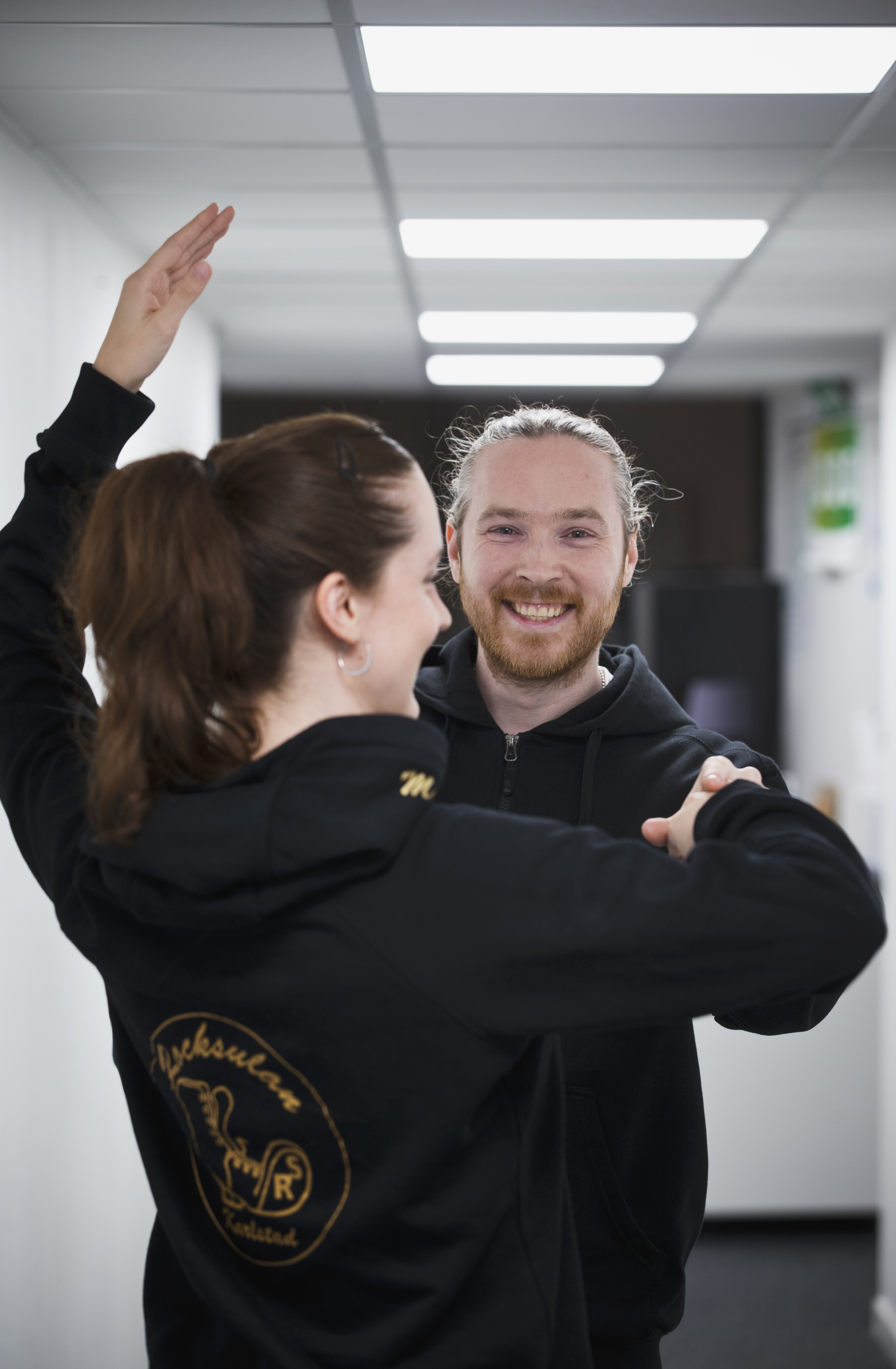 Två personer i svarta hoodies övar en danspose i en hall; kvinnan vänder ryggen till och mannen ler mot kameran.