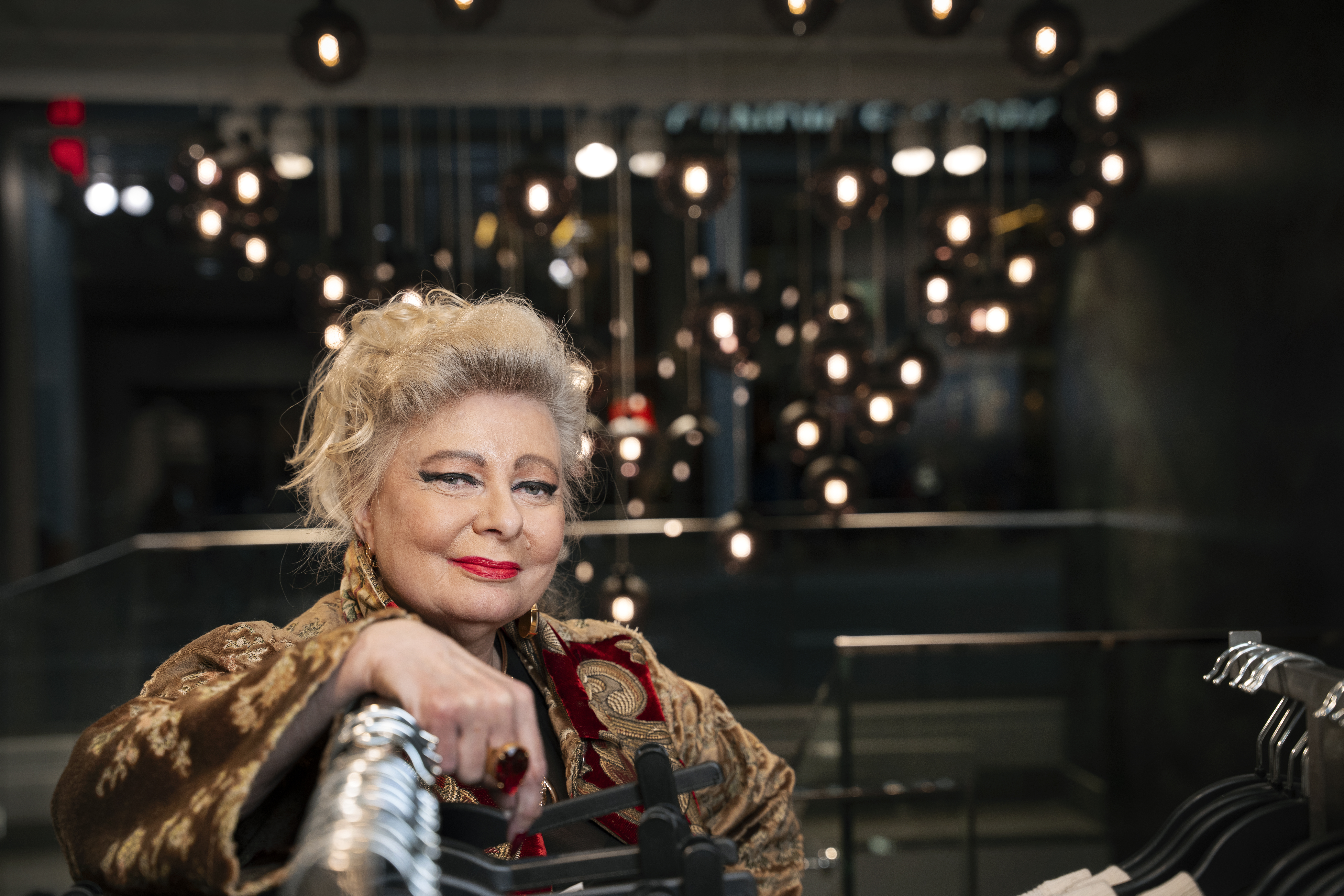 En kvinna med blont hår och rött läppstift står inomhus, lutar sig mot ett klädställ, med många lampor bakom.