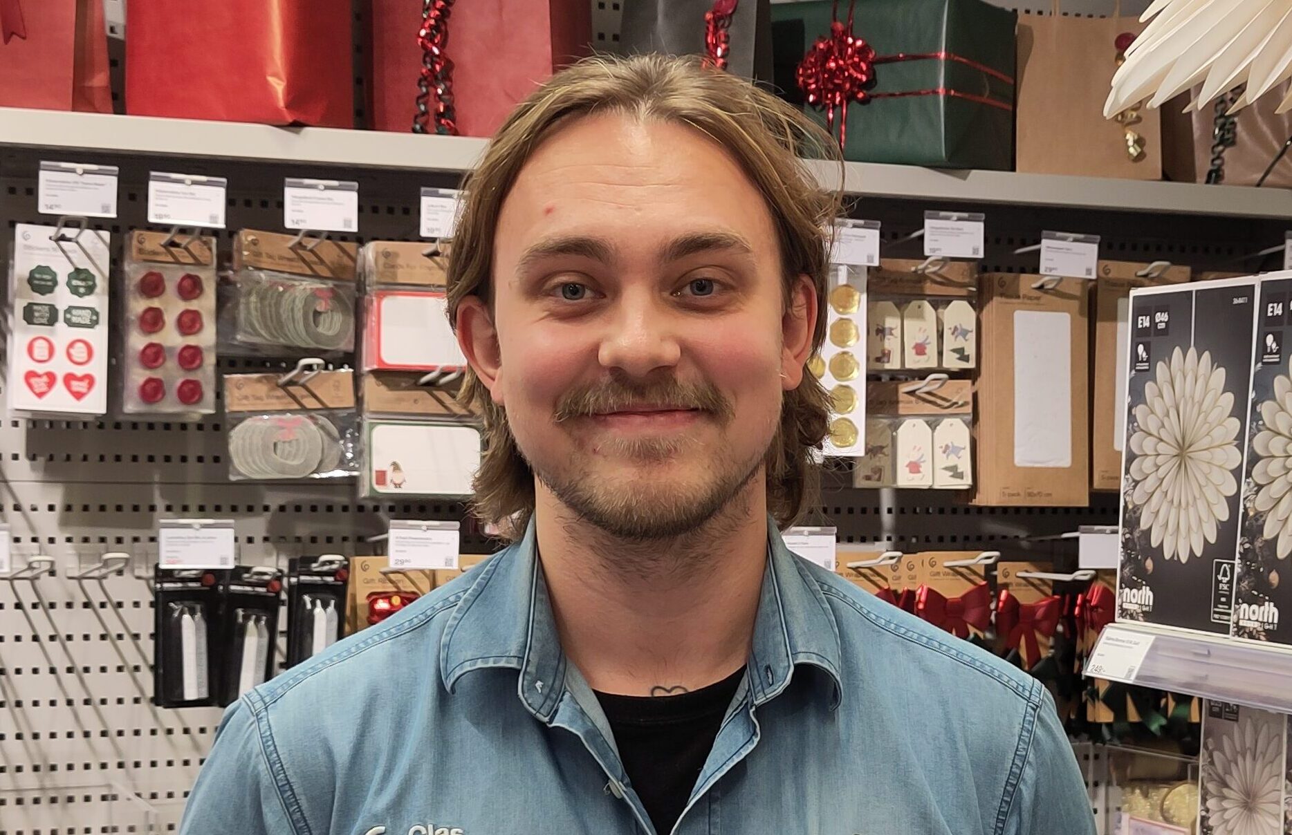 En person med långt hår och mustasch, i blå jeansskjorta, står i en butiksgång med presentpapper och dekorationer på hyllorna bakom.