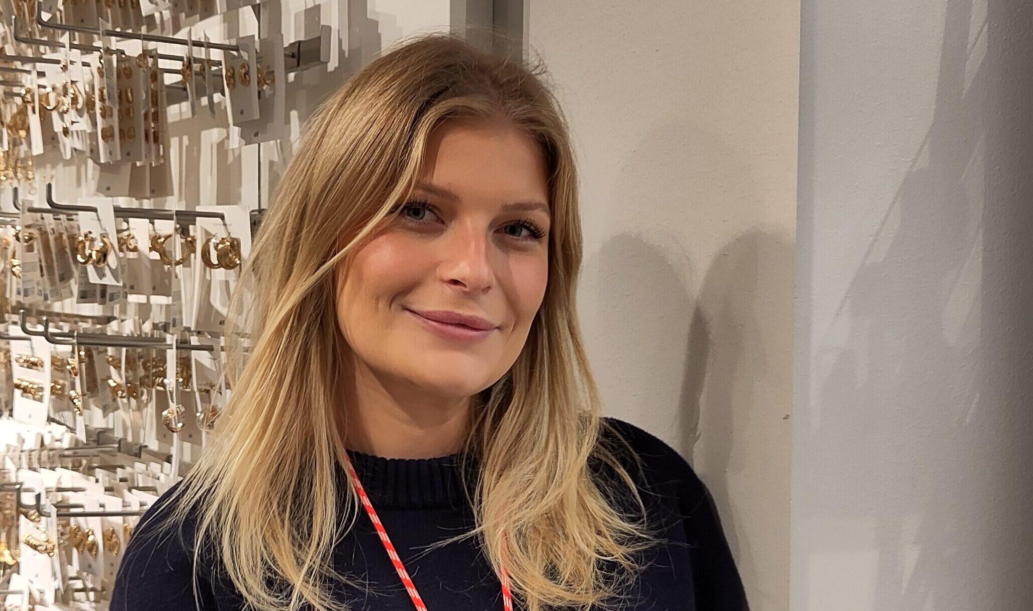 En kvinna med blont hår, mörk tröja och rött nyckelband står framför en vägg med smycken.