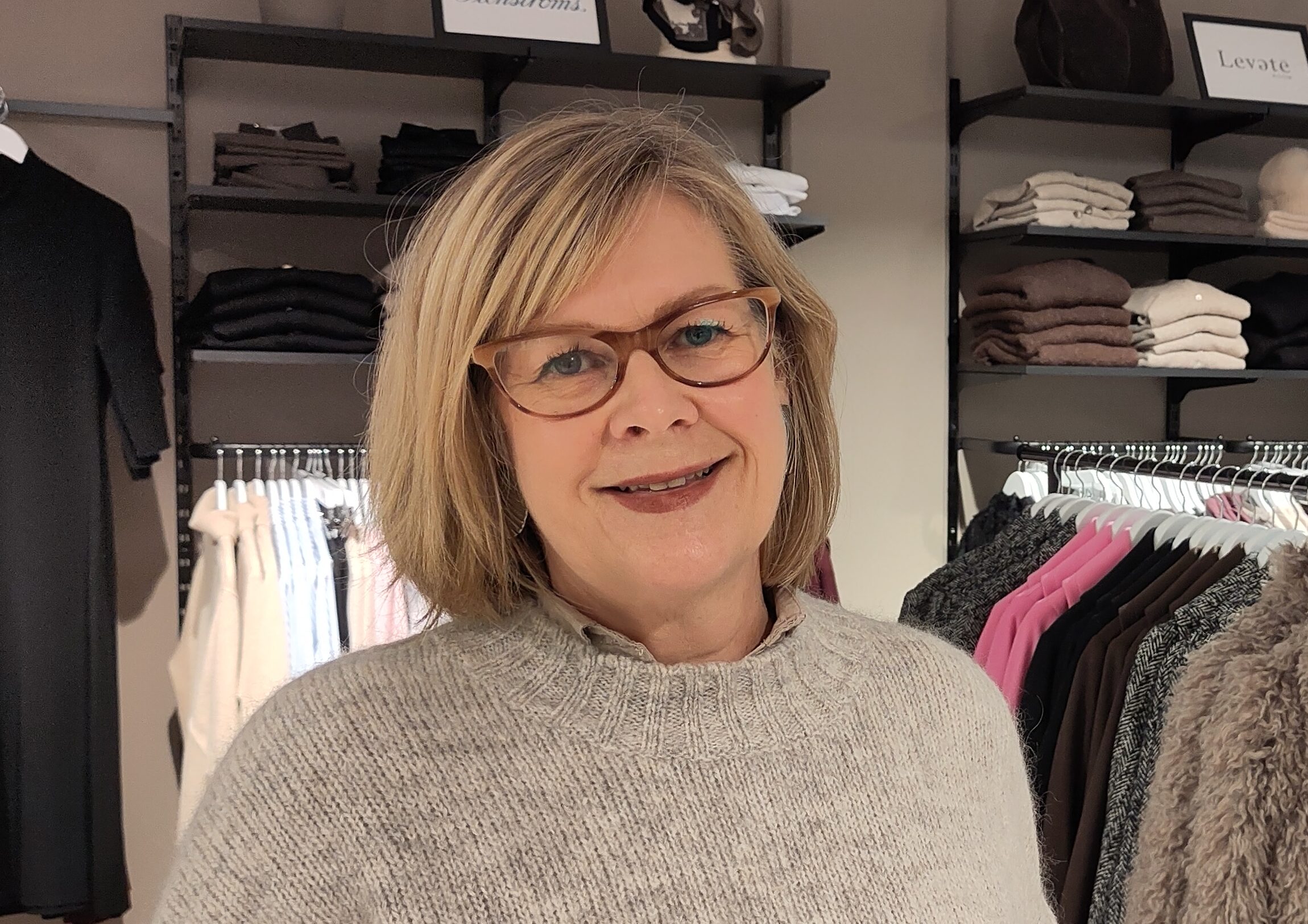 En kvinna med blont hår och glasögon ler i en klädbutik framför vikta tröjor och kläder som hänger.