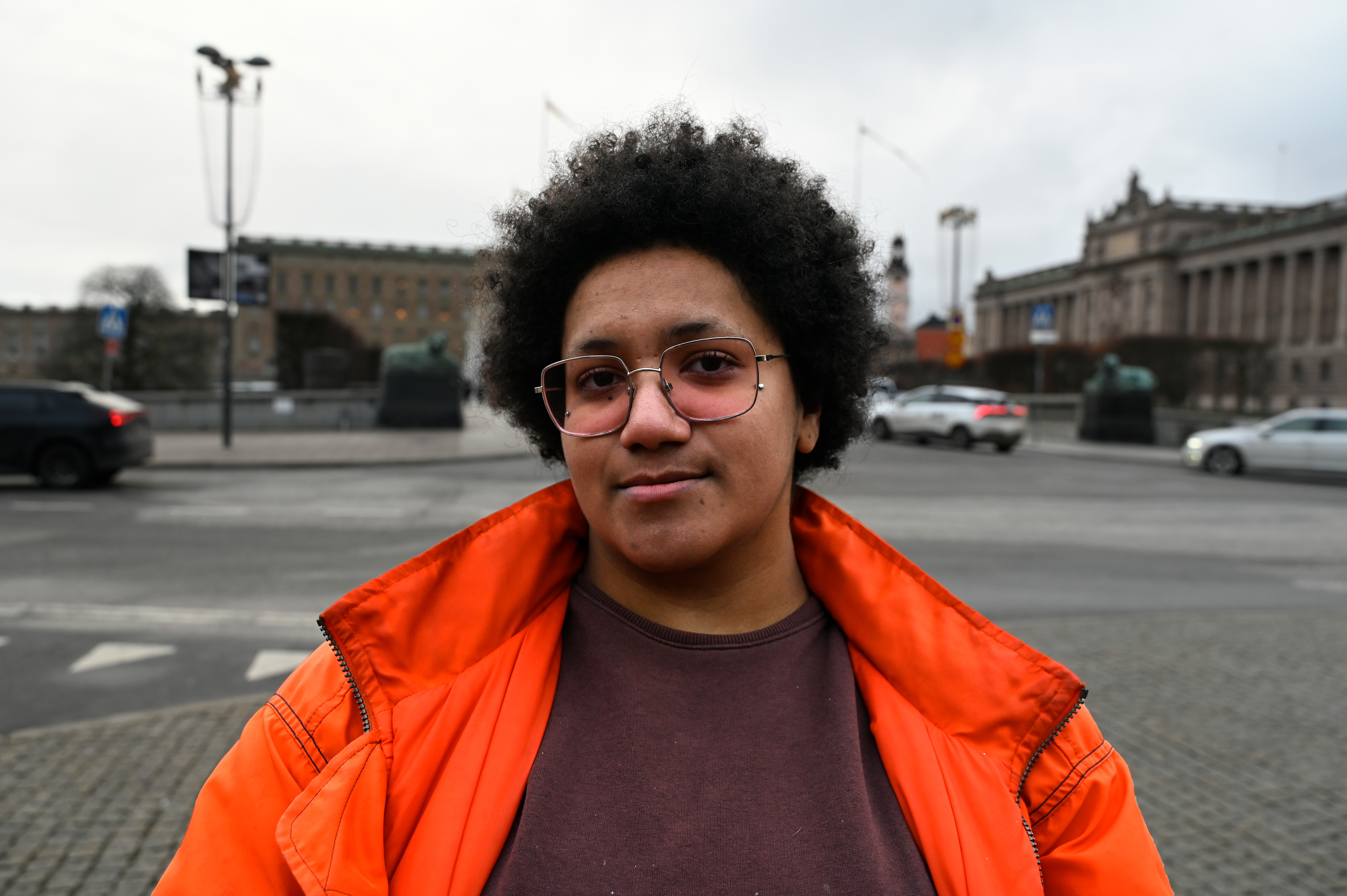 Monica Höglund med orange jacka och rosa glasögon står utomhus på en stadsgata med gamla byggnader i bakgrunden.