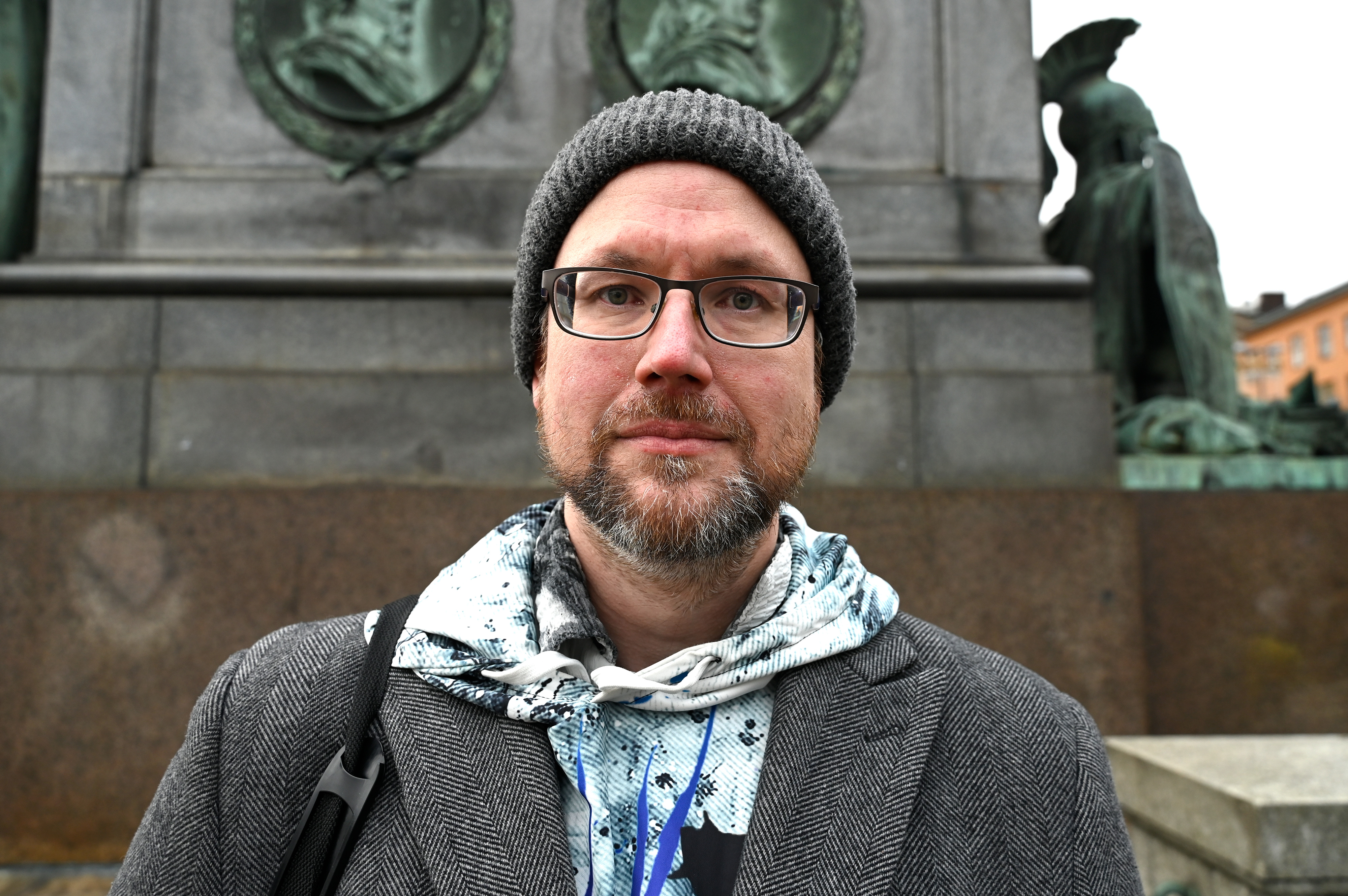 En man med glasögon, grå mössa och mönstrad hoodie står framför en staty med bronsreliefer och figurer utomhus.