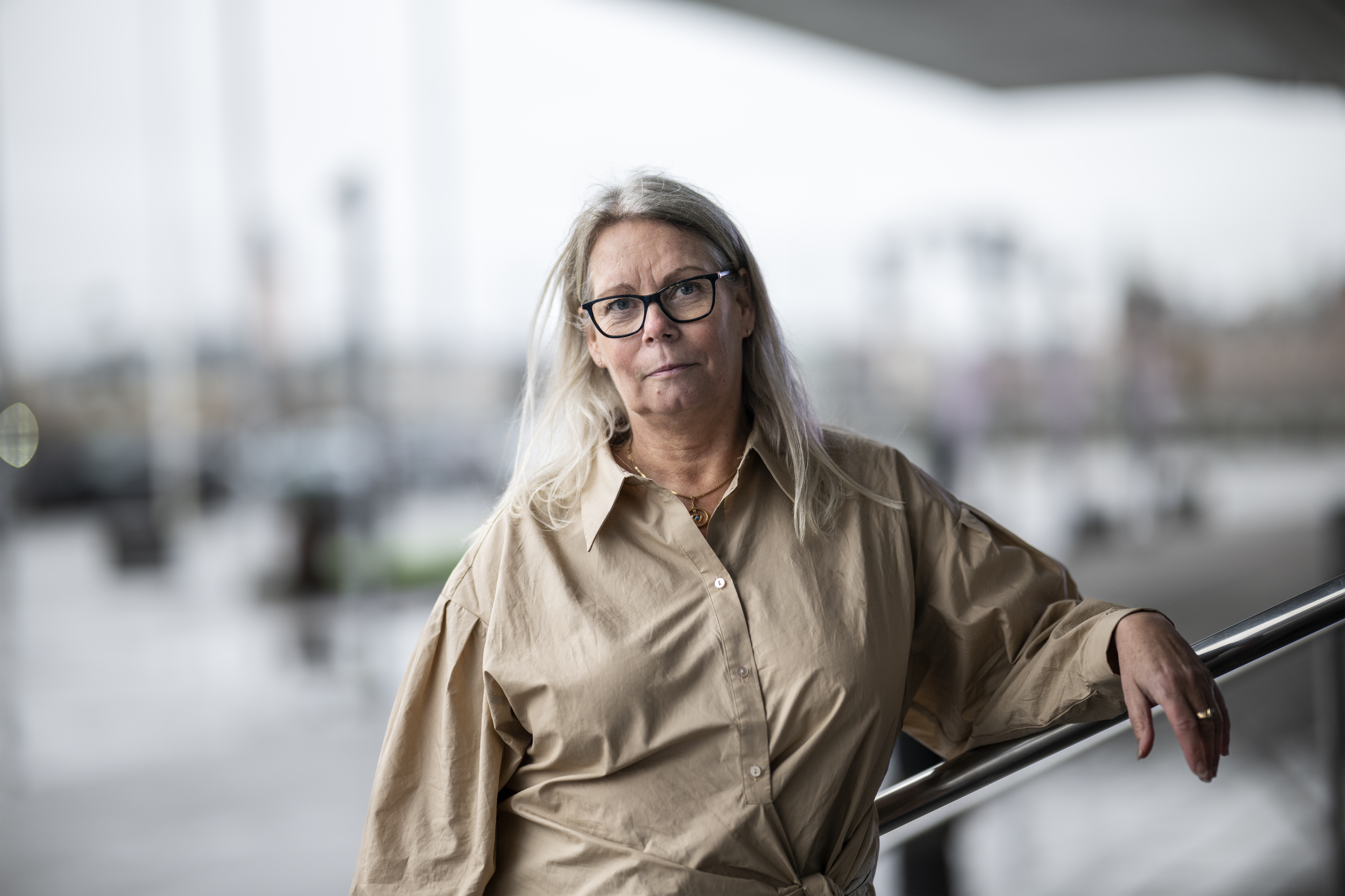 En kvinna med långt grått hår och glasögon står utomhus, lutar sig mot ett räcke, har en beige skjorta på sig, stad i bakgrunden.