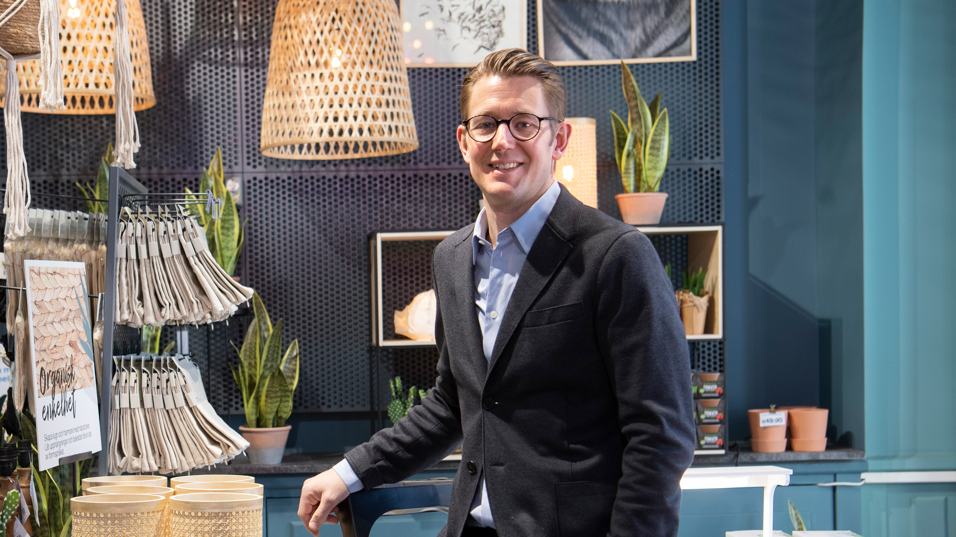 En man med glasögon och mörk kavaj står leende i ett rum med växter, hängande lampor och olika inredningsföremål.