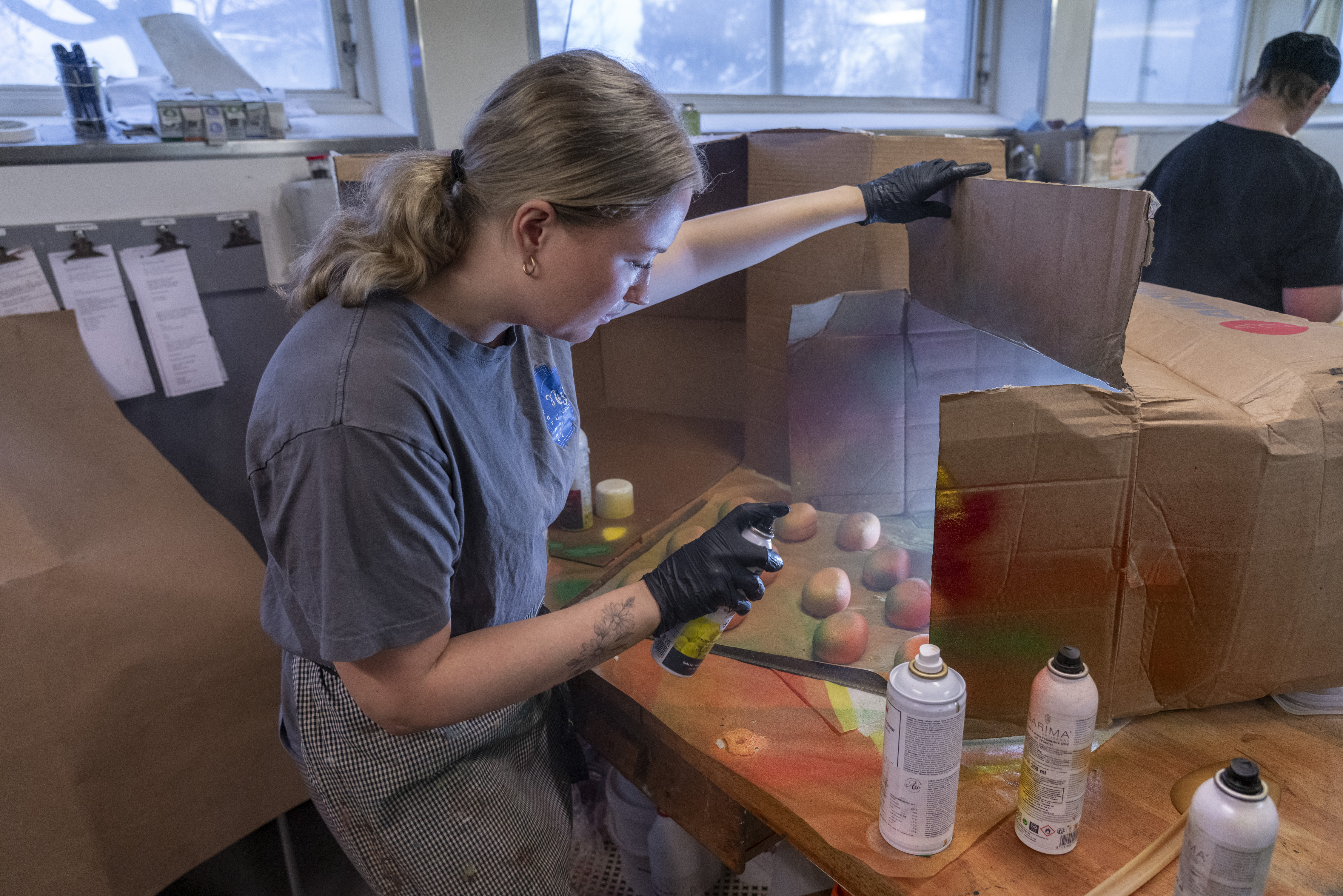 En person med handskar spraymålar konstgjorda päron i en stor kartong på ett arbetsbord, burkar med sprayfärg står bredvid.