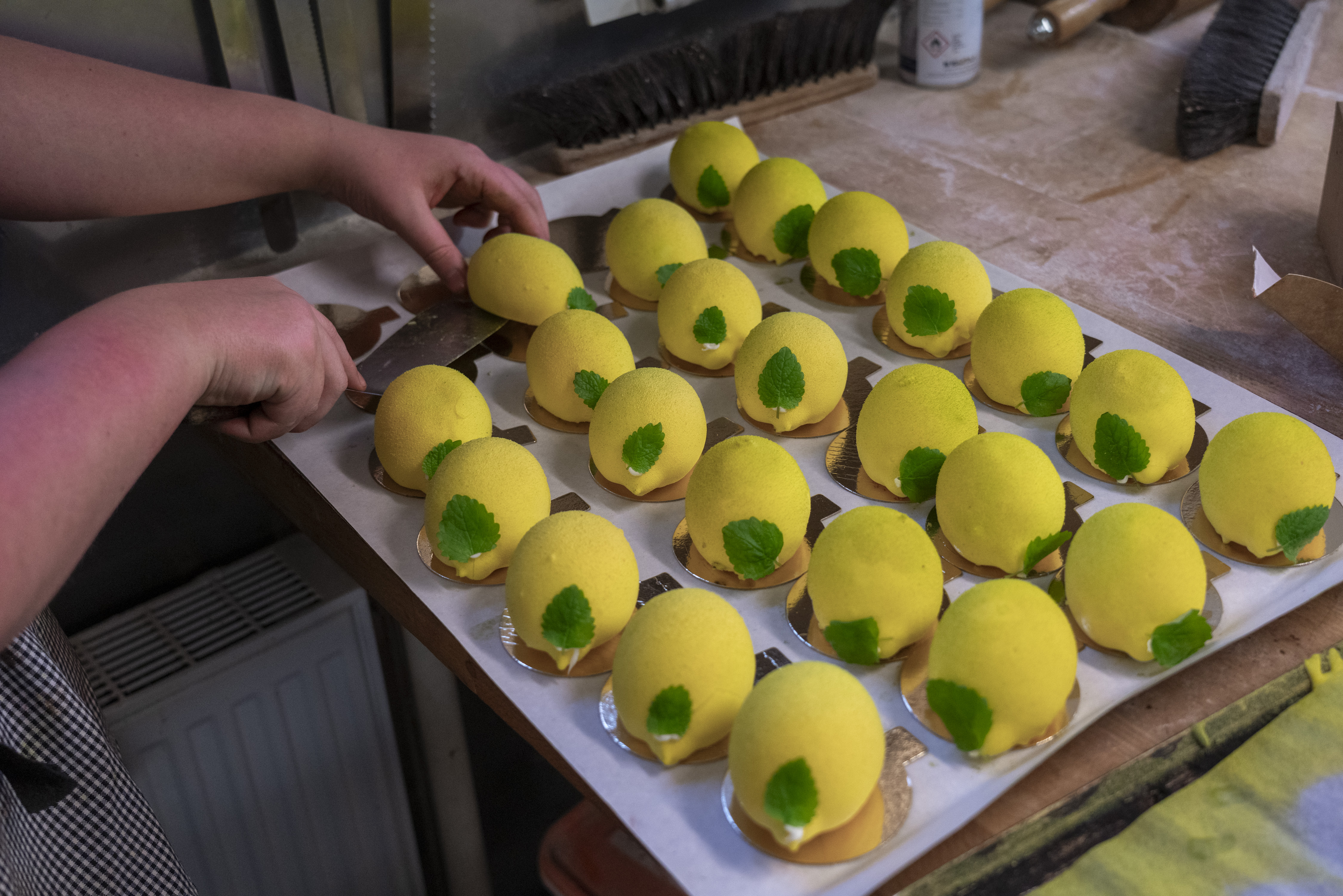 En person placerar flera gula, citronformade bakverk med gröna bladdekorationer på en bricka i ett kök.