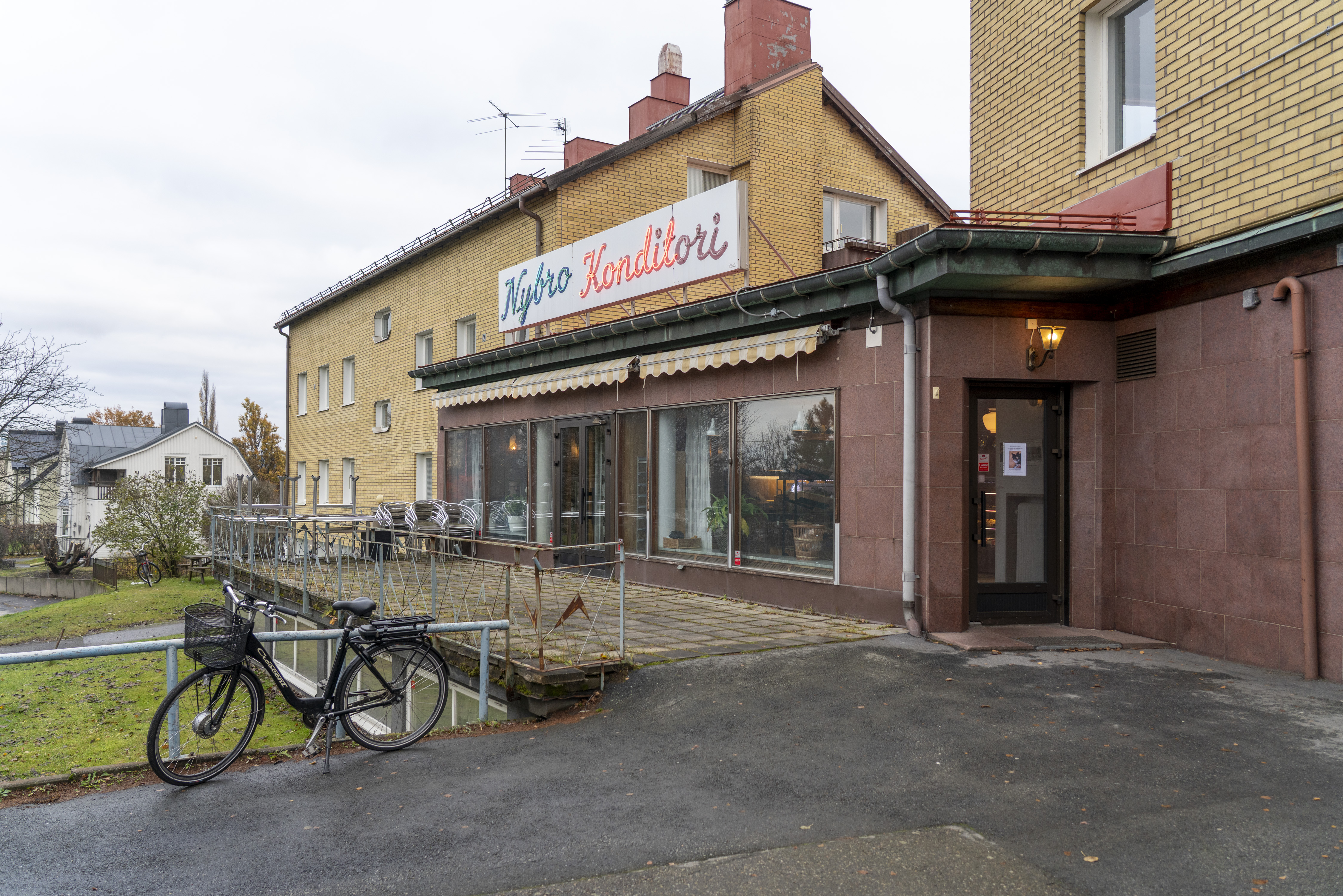 En byggnad av tegel med stora fönster och en skylt där det står "Nybro Konditori". En cykel står nära på trottoaren.