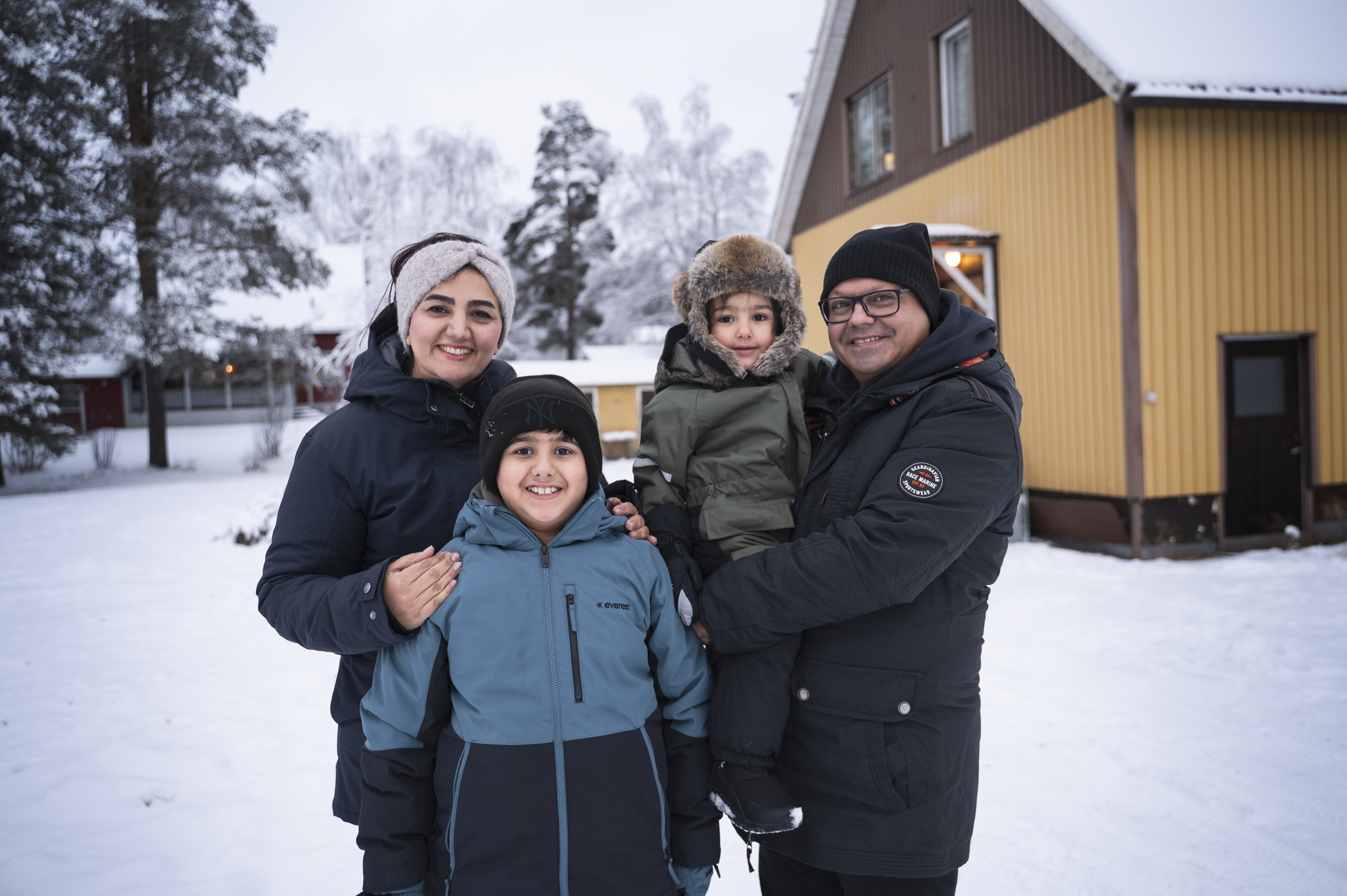 En familj på fyra med vinterkläder står utanför ett gult hus i snön och ler mot kameran.