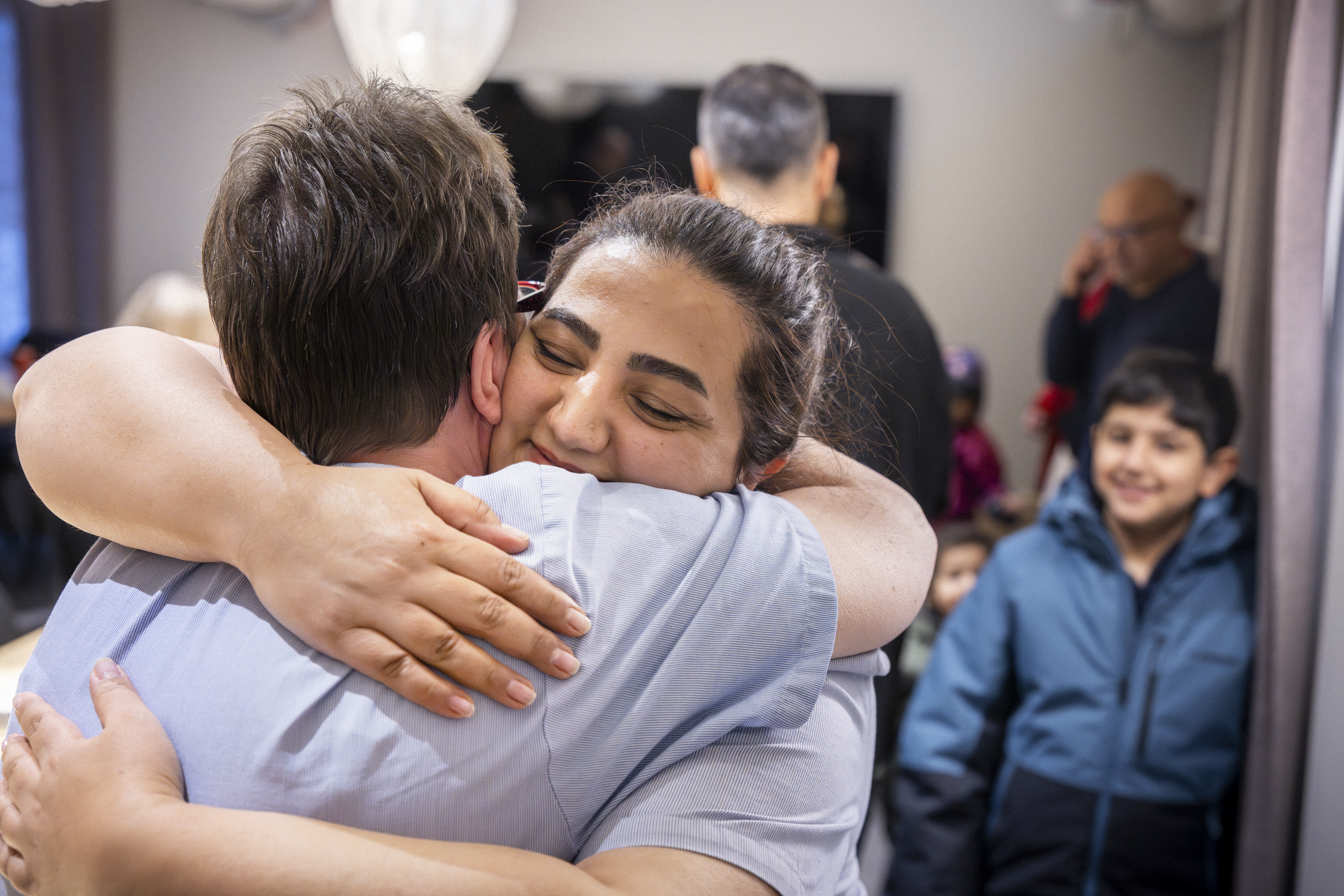 Två personer kramas i förgrunden, omgivna av flera andra i en inomhusmiljö med stark belysning.