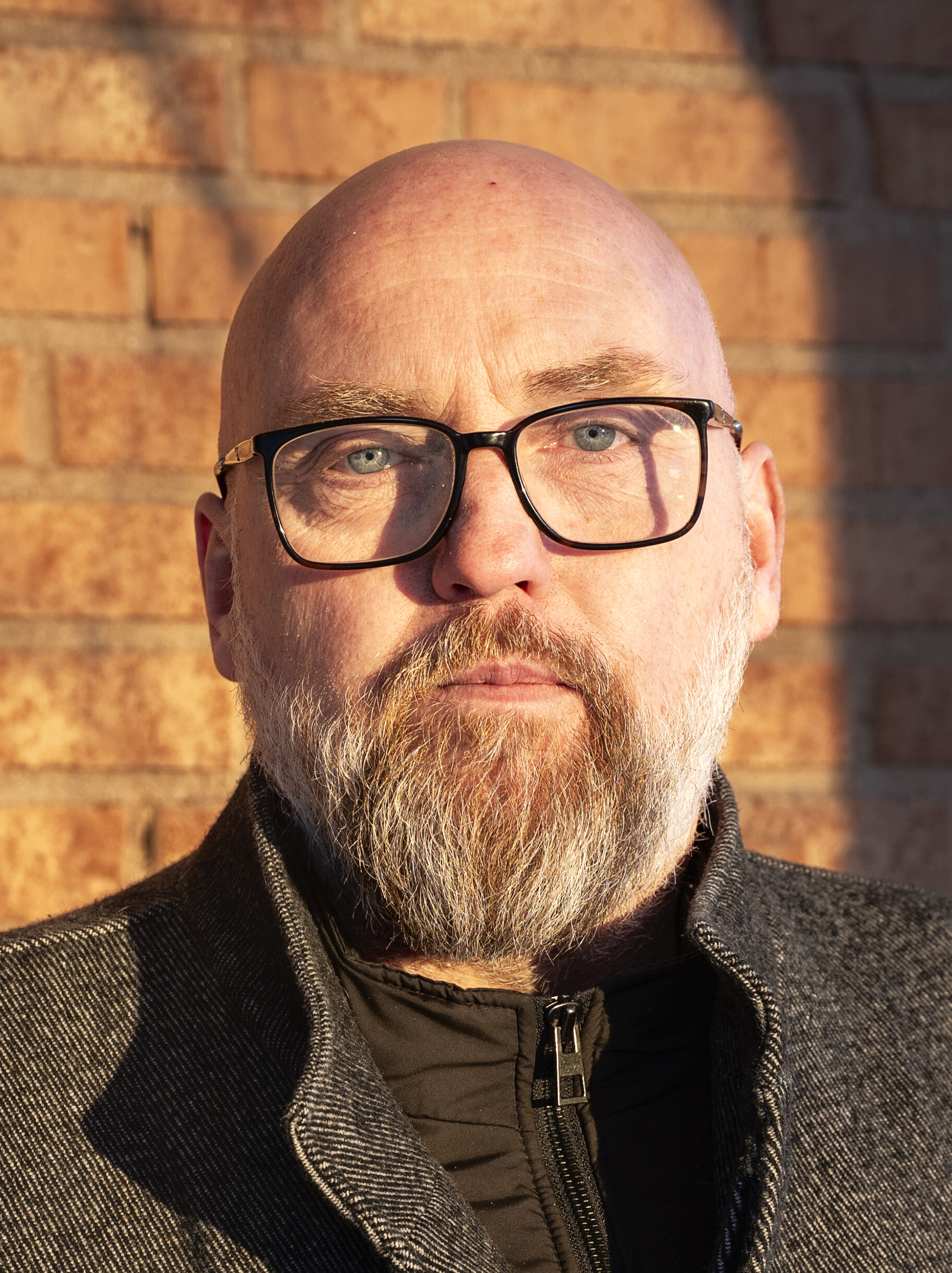 Jan Lindberg med glasögon och skägg står mot en tegelvägg i solskenet och kastar en skugga.