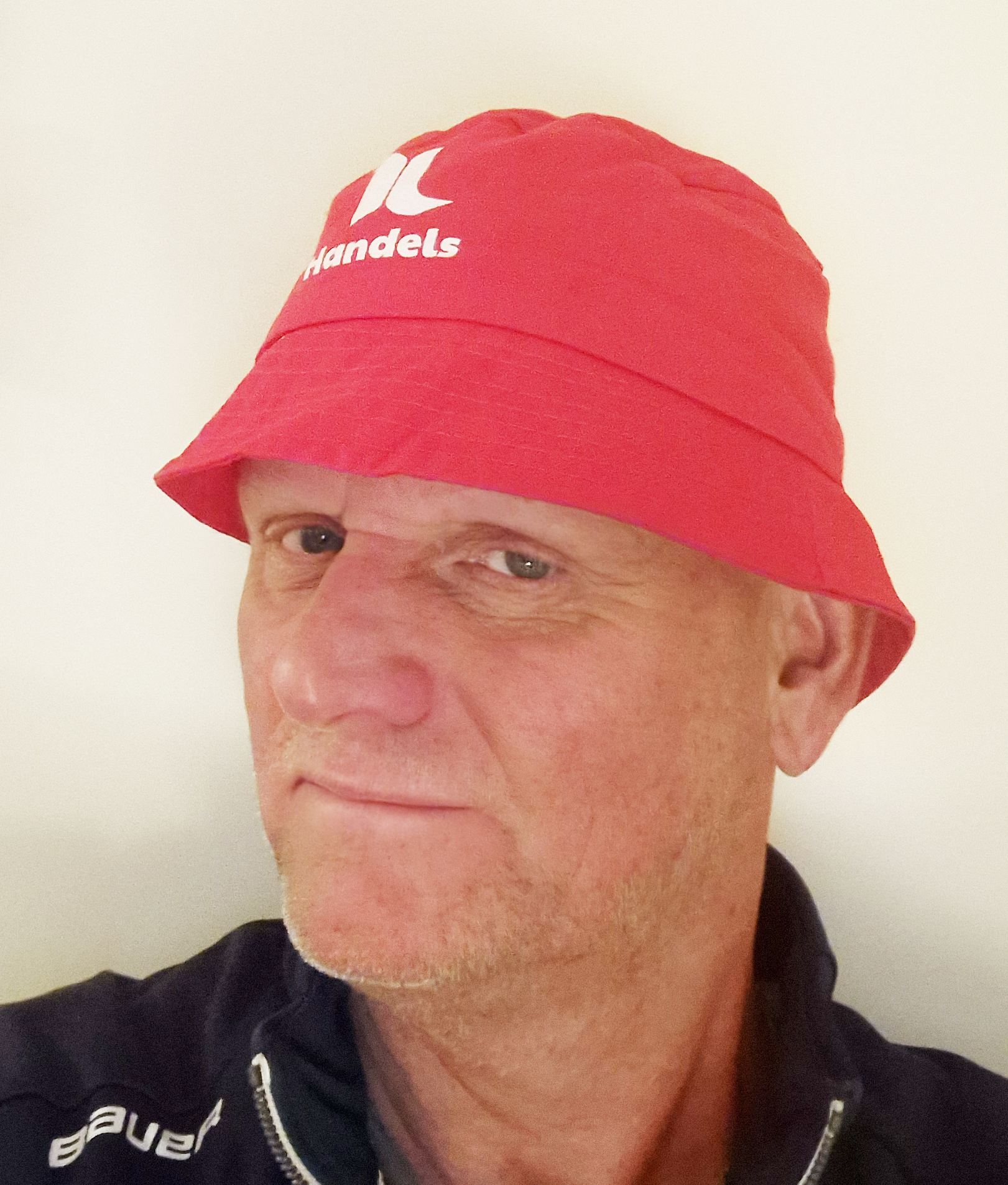 En man med röd hatt med vit logotyp och mörk zip-jacka, tittar lite åt sidan mot en enfärgad bakgrund.