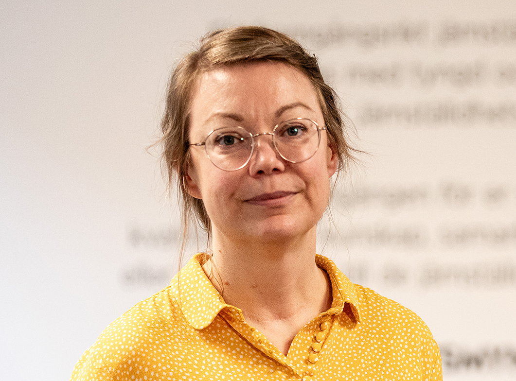 En kvinna med glasögon och ljusbrunt bakåtdraget hår har på sig en gulprickig skjorta och tittar mot kameran.