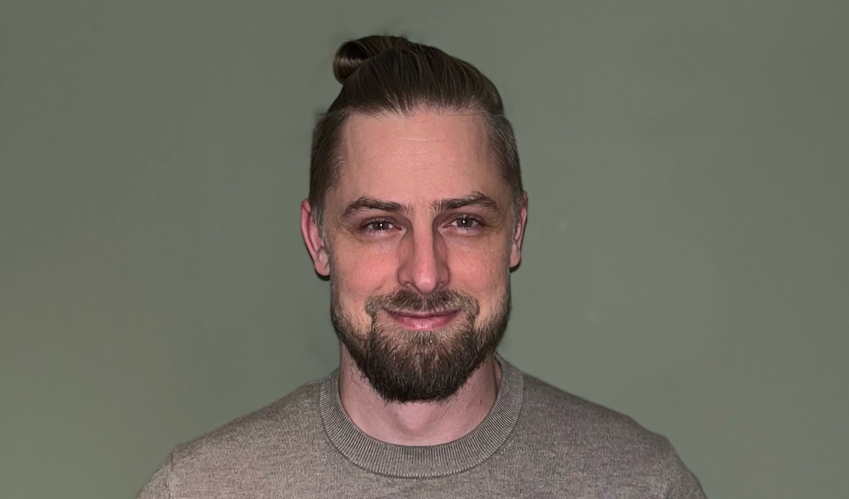 En man med kort skägg och mustasch, iförd en grå tröja, ler framför en enfärgad grön bakgrund.